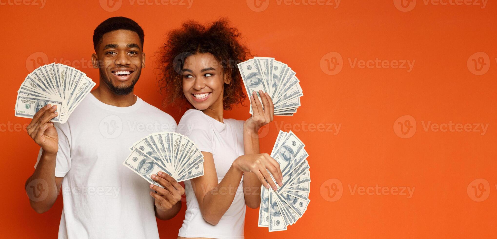 Happy millennial couple posing with lot of money, holding with dollar fans, orange panorama background with empty space photo