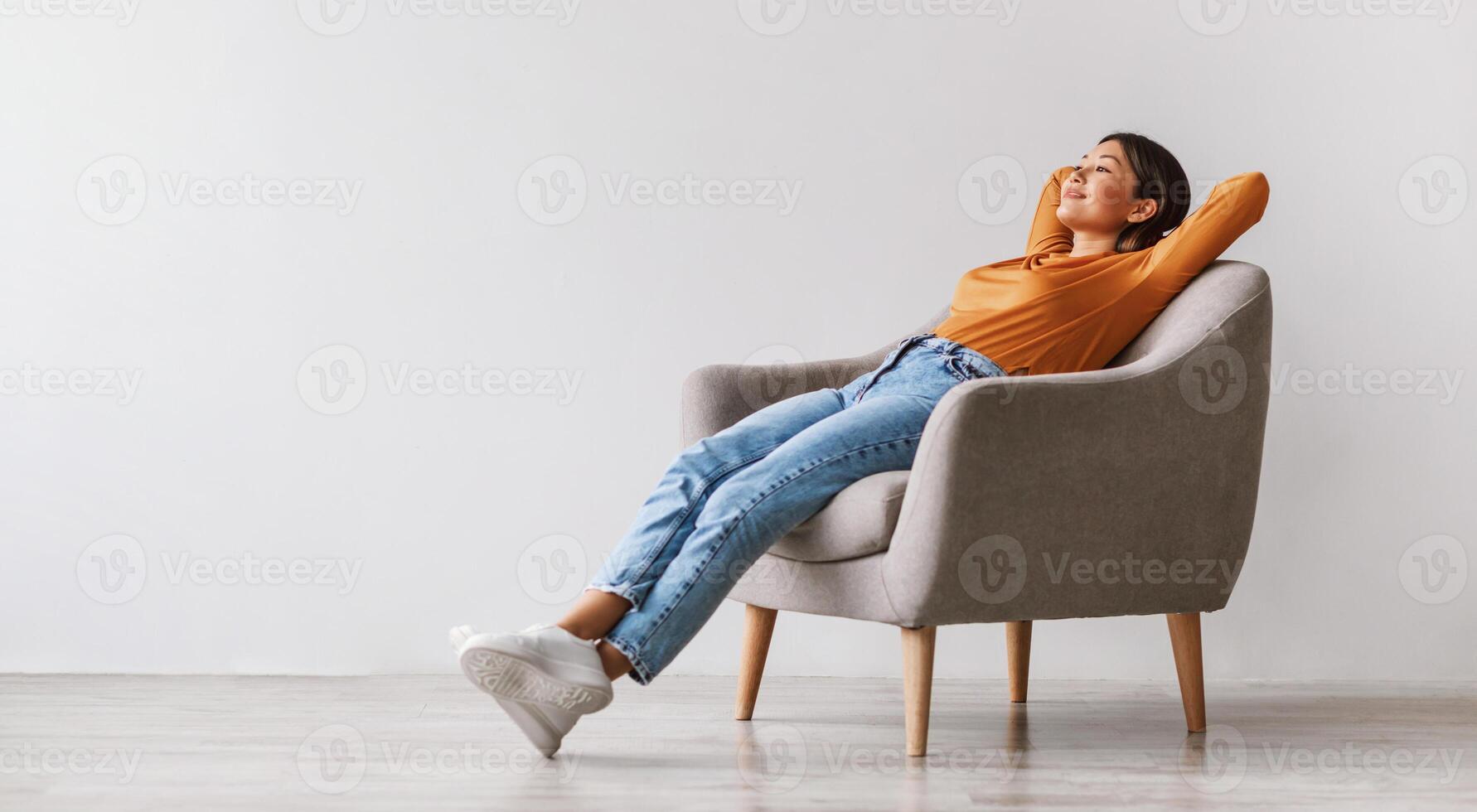 Gorgeous Asian woman sitting in comfy armchair, being lazy, feeling peaceful against white studio wall, panorama with copy space. Attractive young lady resting and relaxing with hands behind head photo