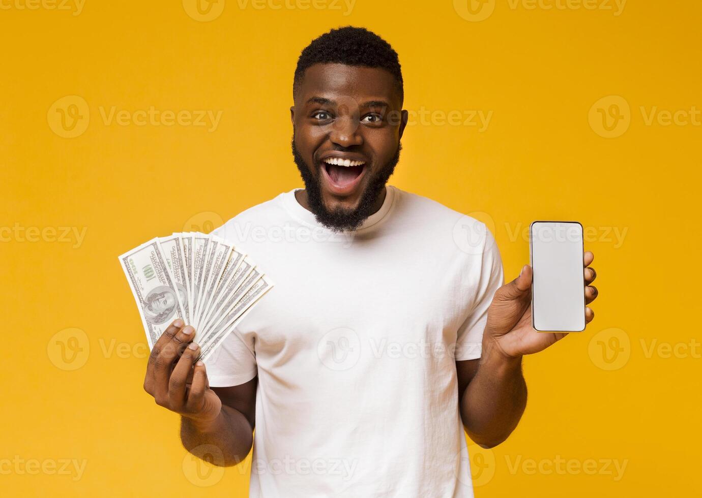 Making money on internet. Happy african-american guy showing showing cellphone with blank screen and bunch of money, orange background, free space photo