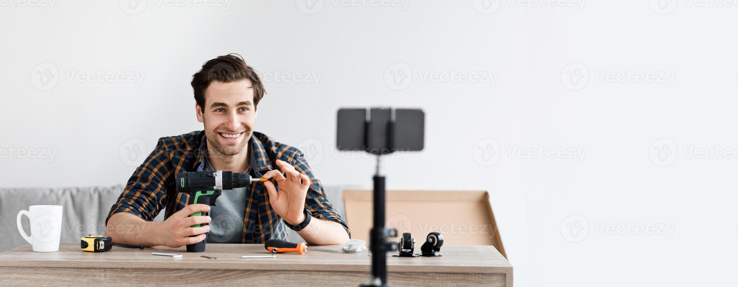 Carpenter recording for repair blog or review of instruments. Happy attractive millennial man looks at smartphone in webcam and shows electric screwdriver, in living room interior with tools photo