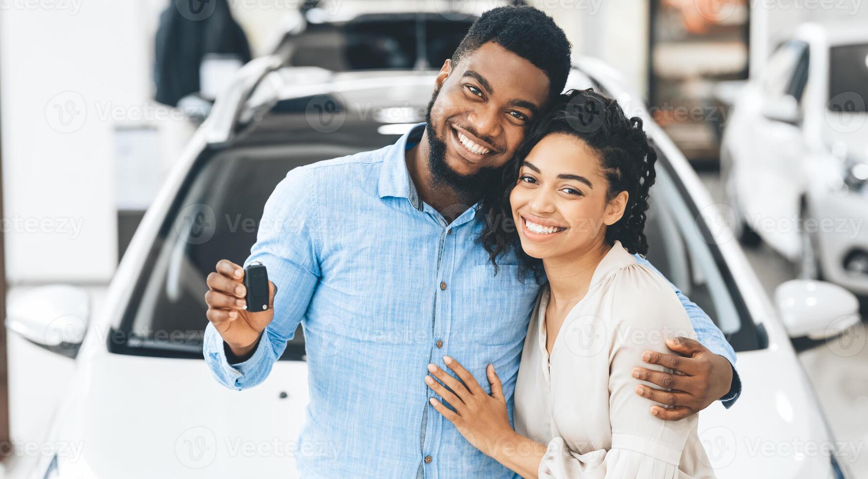Black Family Couple Showing New Car Key Smiling To Camera In Dealership Showroom. Happy Spouses Hugging Near Their Auto. Panorama photo