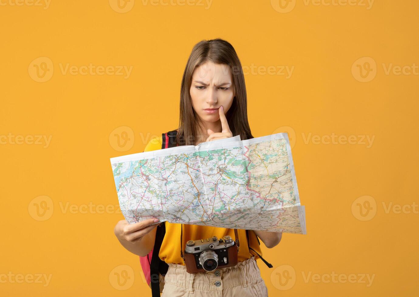 Confused woman traveler with camera looking at city map, got lost, yellow studio background, copy space. Upset young lady backpacker lost in city, checking map. Navigation, trekking during travelling photo