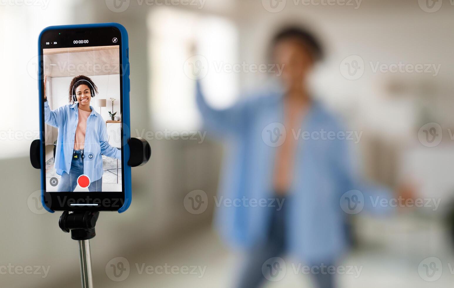 Modern Content Creator. Positive African American young lady in headset filming blog for social network, using cellphone on tripod for broadcast in living room, selective focus on device screen photo