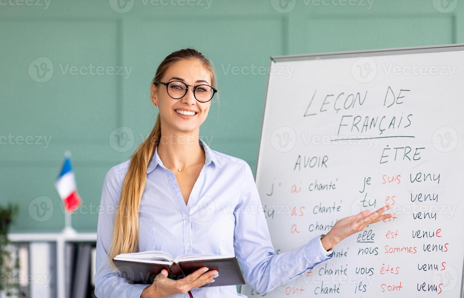 Happy French teacher explaining foreign language rules near blackboard indoors and smiling at camera. Confident young female tutor teaching linguistic online course photo