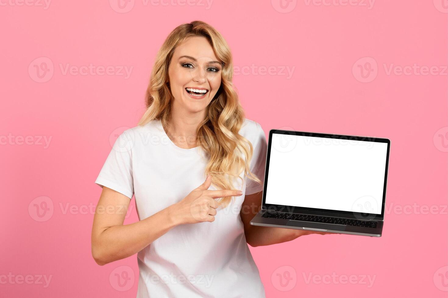 A cheerful young woman with long blonde hair happily points to a blank laptop screen while standing against a bright pink background, highlighting the device's empty display. photo