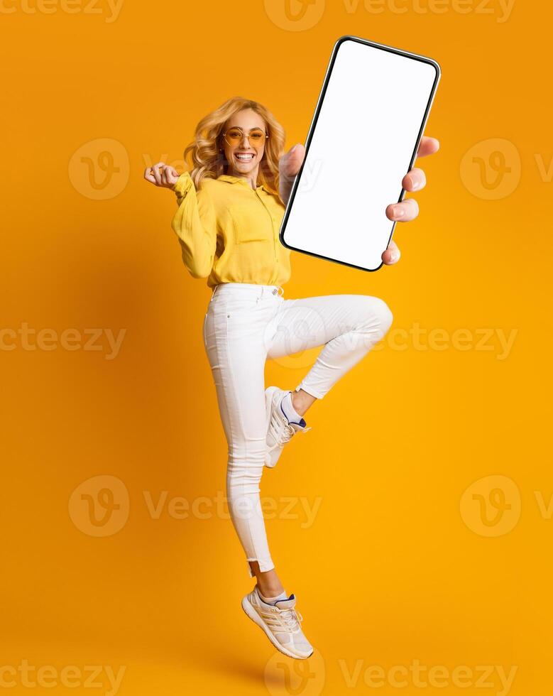 Cheerful blonde woman jumping up and showing newest smartphone with empty screen, enjoying new application for mobile device. Orange studio background, creative collage with mockup photo