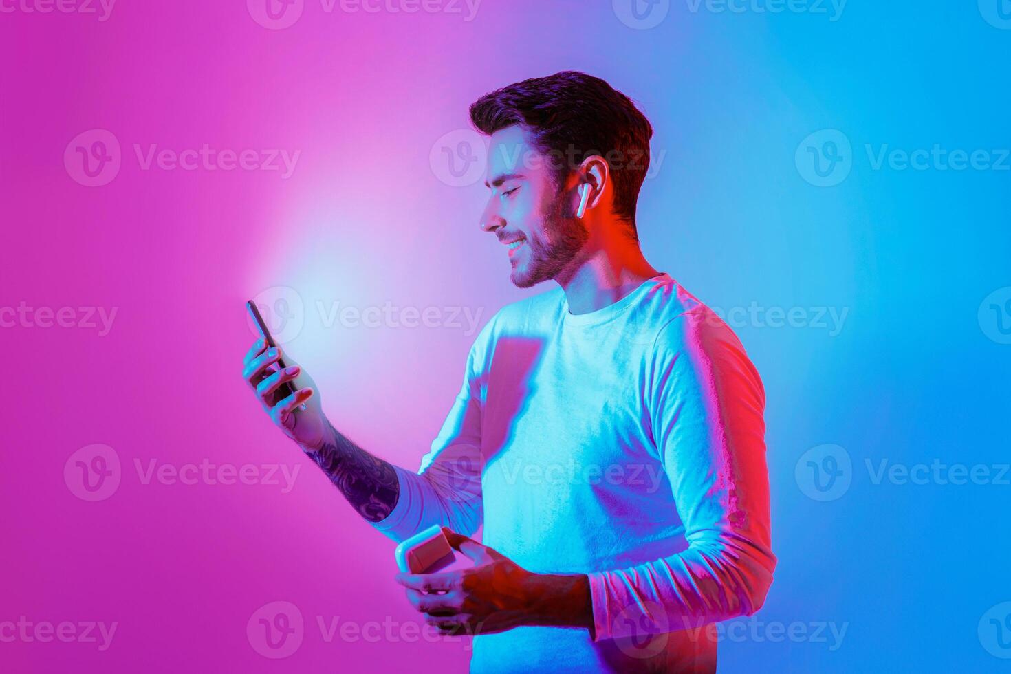 Watching . Smiling millennial guy with wireless headphones, looks at smartphone with luminous screen, in neon, studio shot photo