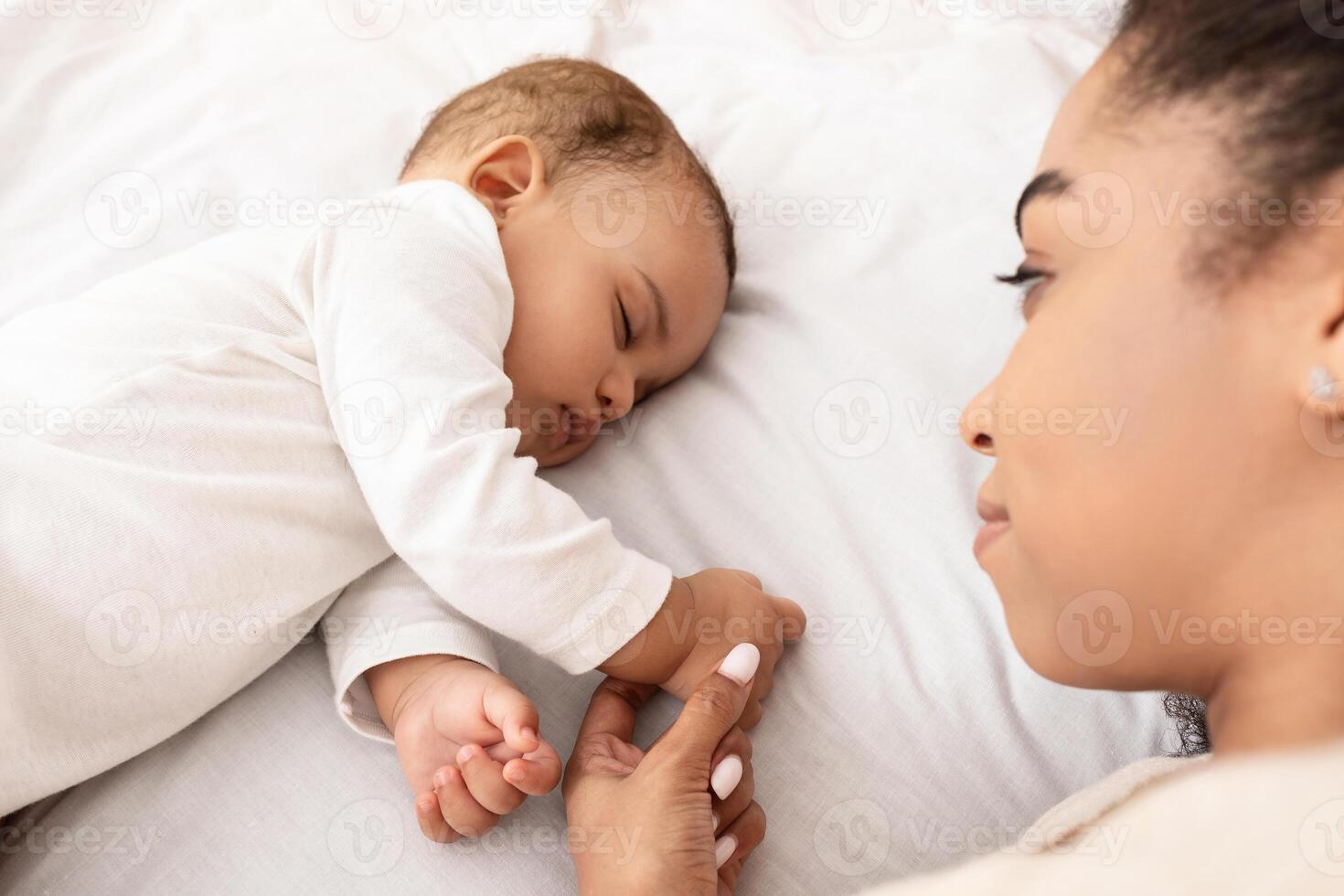 vista desde arriba de dormido africano americano bebé infantil y su madre acostado en cama en dormitorio a hogar. mamá participación su pequeño niñito hijos mano mientras él duerme pacíficamente selectivo atención foto