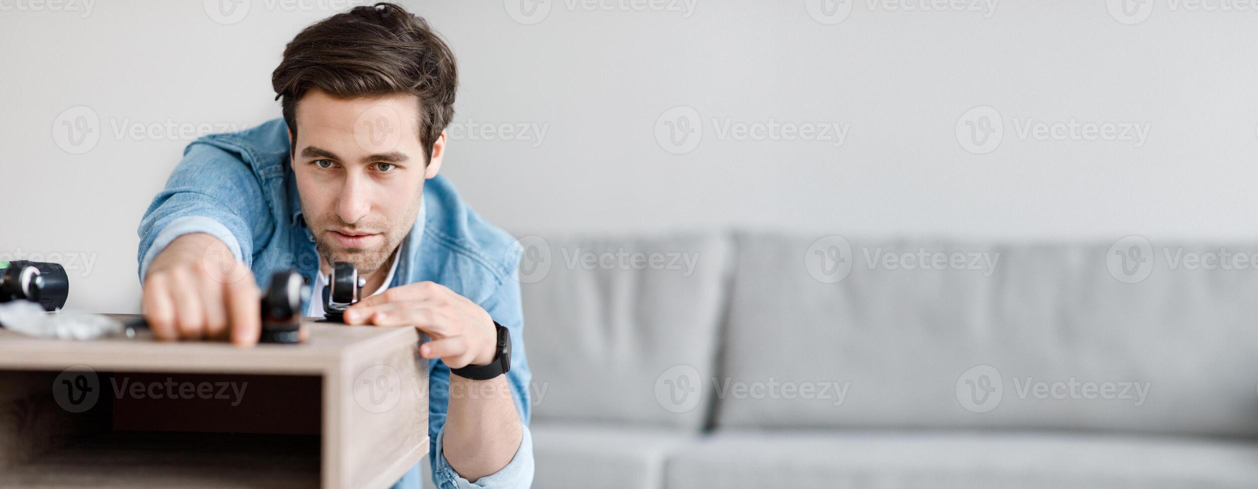 Job from home, carpentry, handyman collects desk and work, new online ordering and hobby. Busy young handsome guy assembles furniture in living room interior, empty space photo