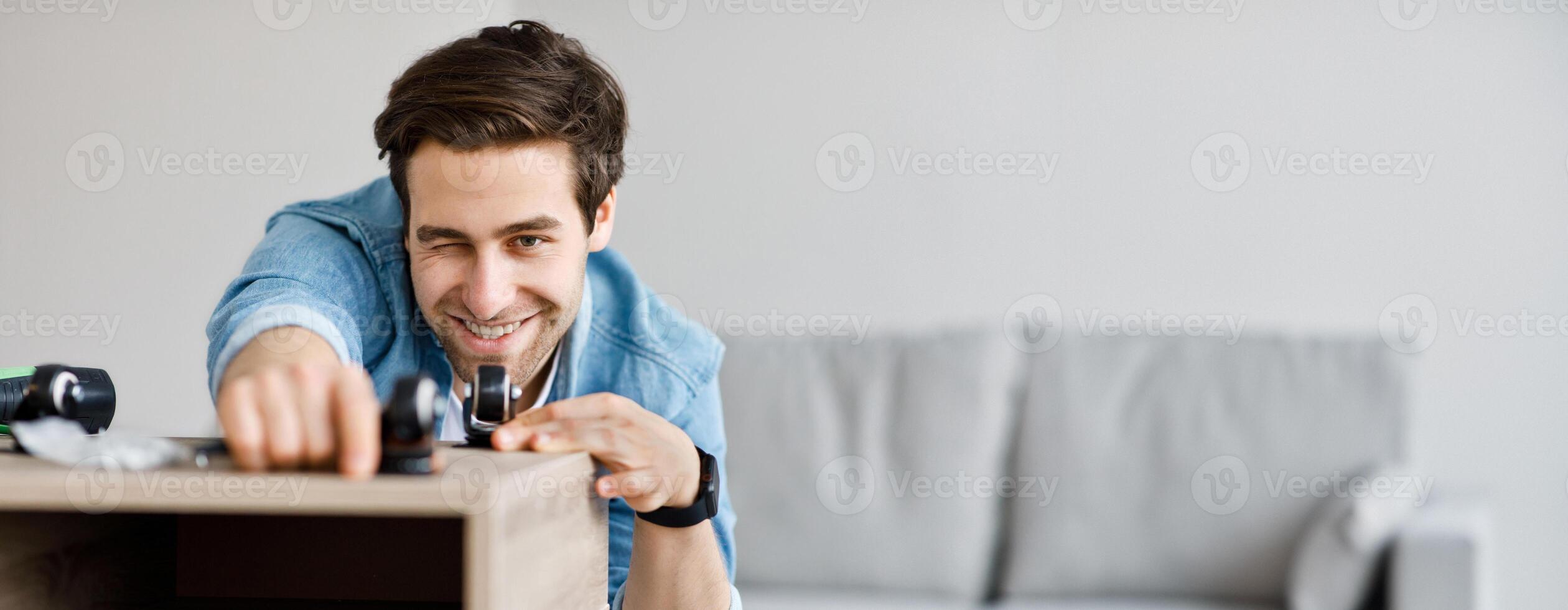 Self made modern furniture and hobby, engineer with tools, homemade decoration. Young smiling man assembling wooden bookshelf or desk measures distance between wheels on table in living room interior photo
