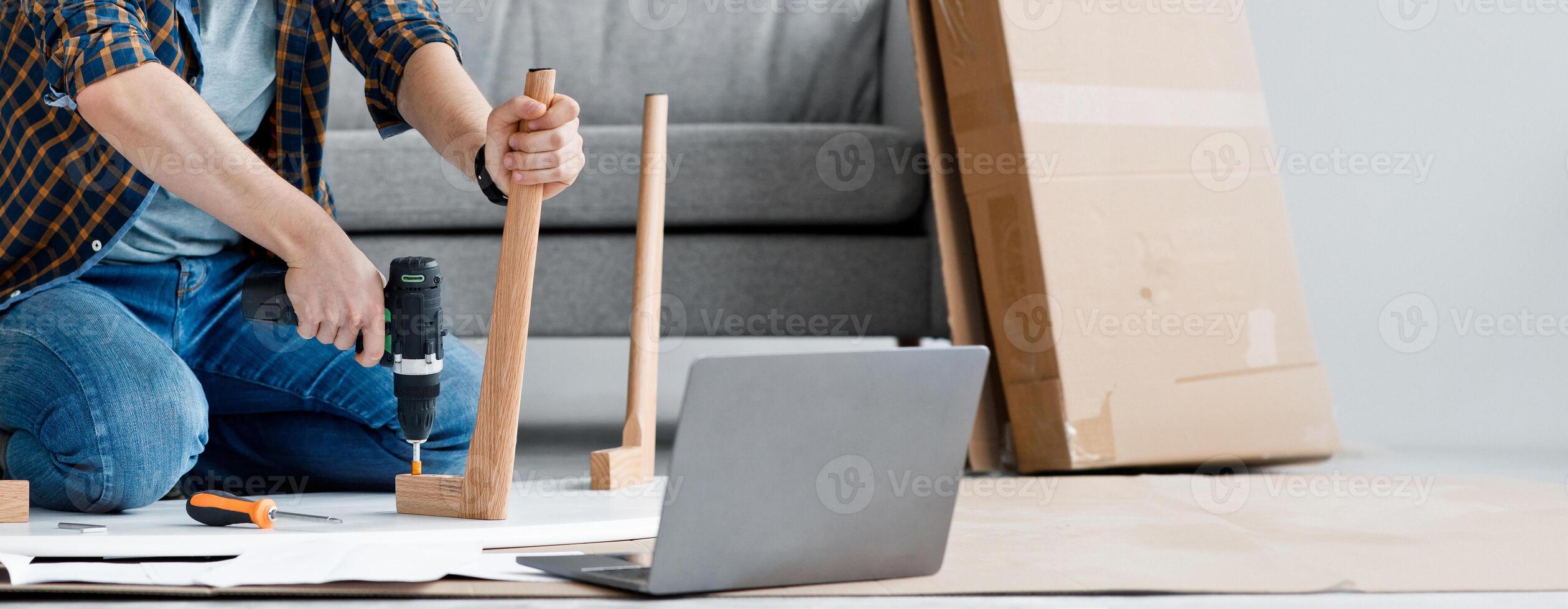 Furnishing house. Millennial guy fixing furniture, repairing wooden table uses electric drill, sits on floor at home, screwing detail with screwdriver and looks at laptop in living room interior photo