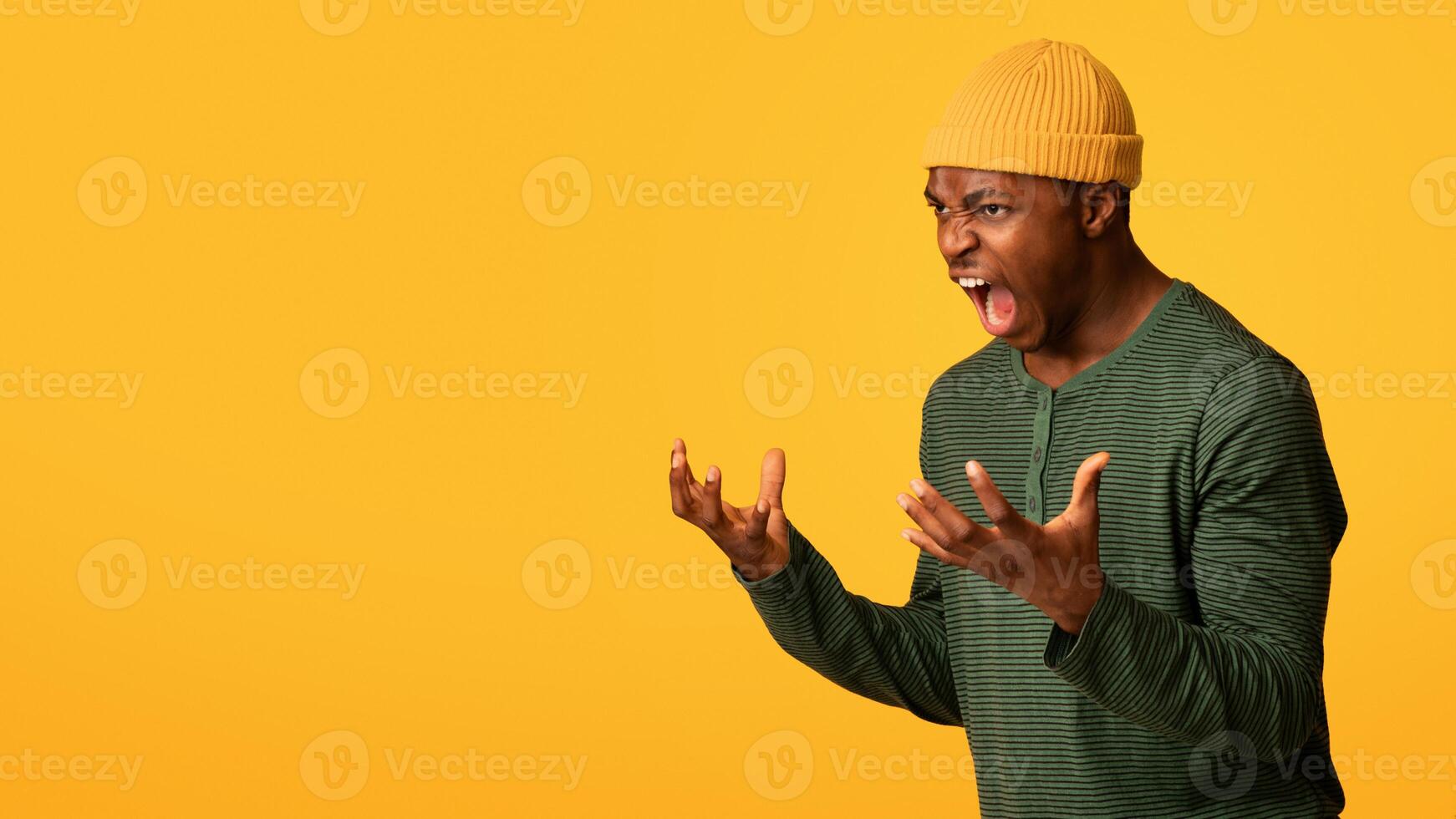 Portrait Of Angry African American Man Screaming With Rage, Furious Agressive Young Black Guy Raising Hands And Shouting While Standing Over Yellow Background In Studio, Panorama With Copy Space photo