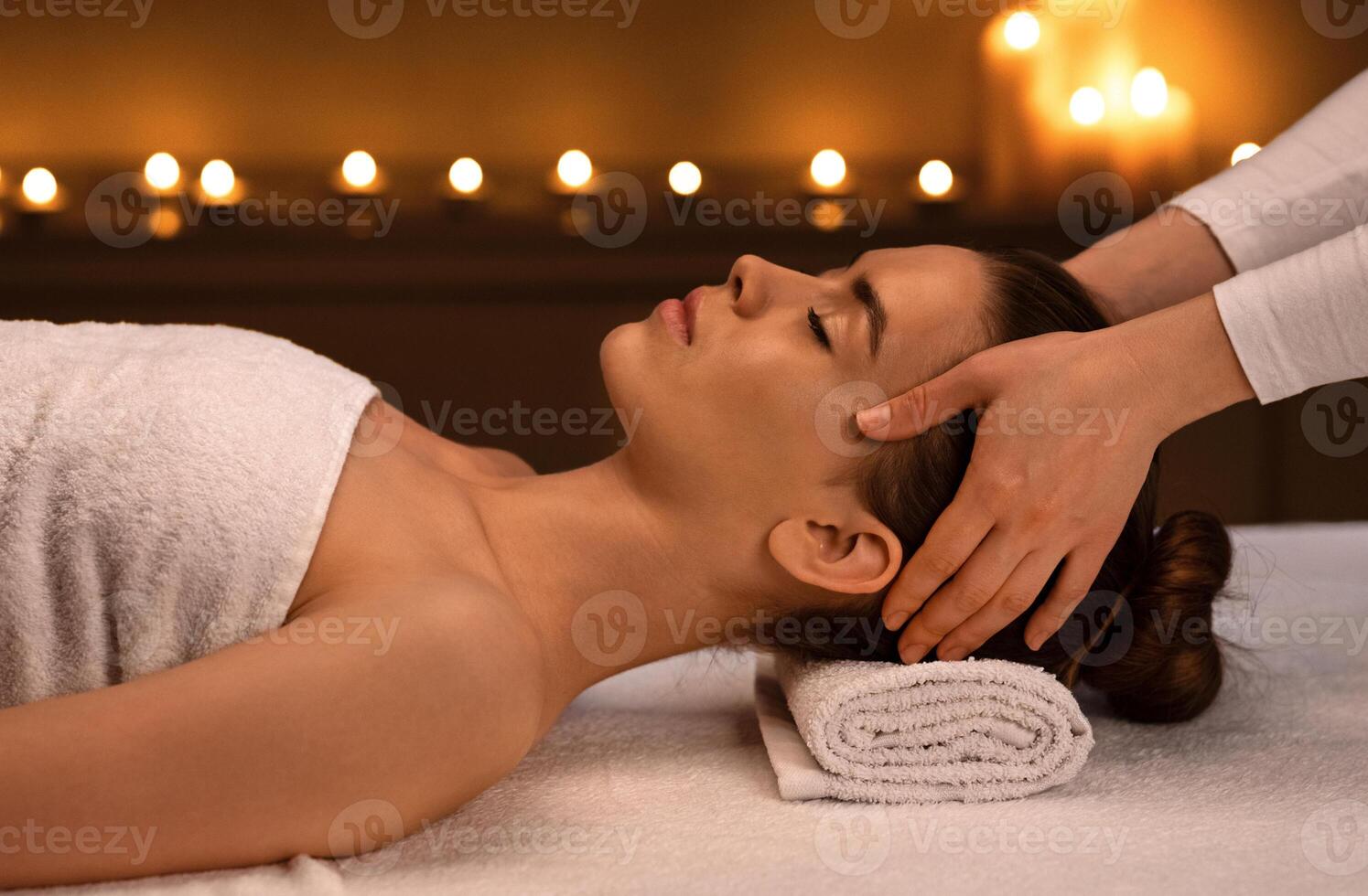 Joyful young woman getting acupressure massage at modern luxury spa in romantic atmosphere, side view. Spa therapist making healing massage for young beautiful lady, pressing on her head photo