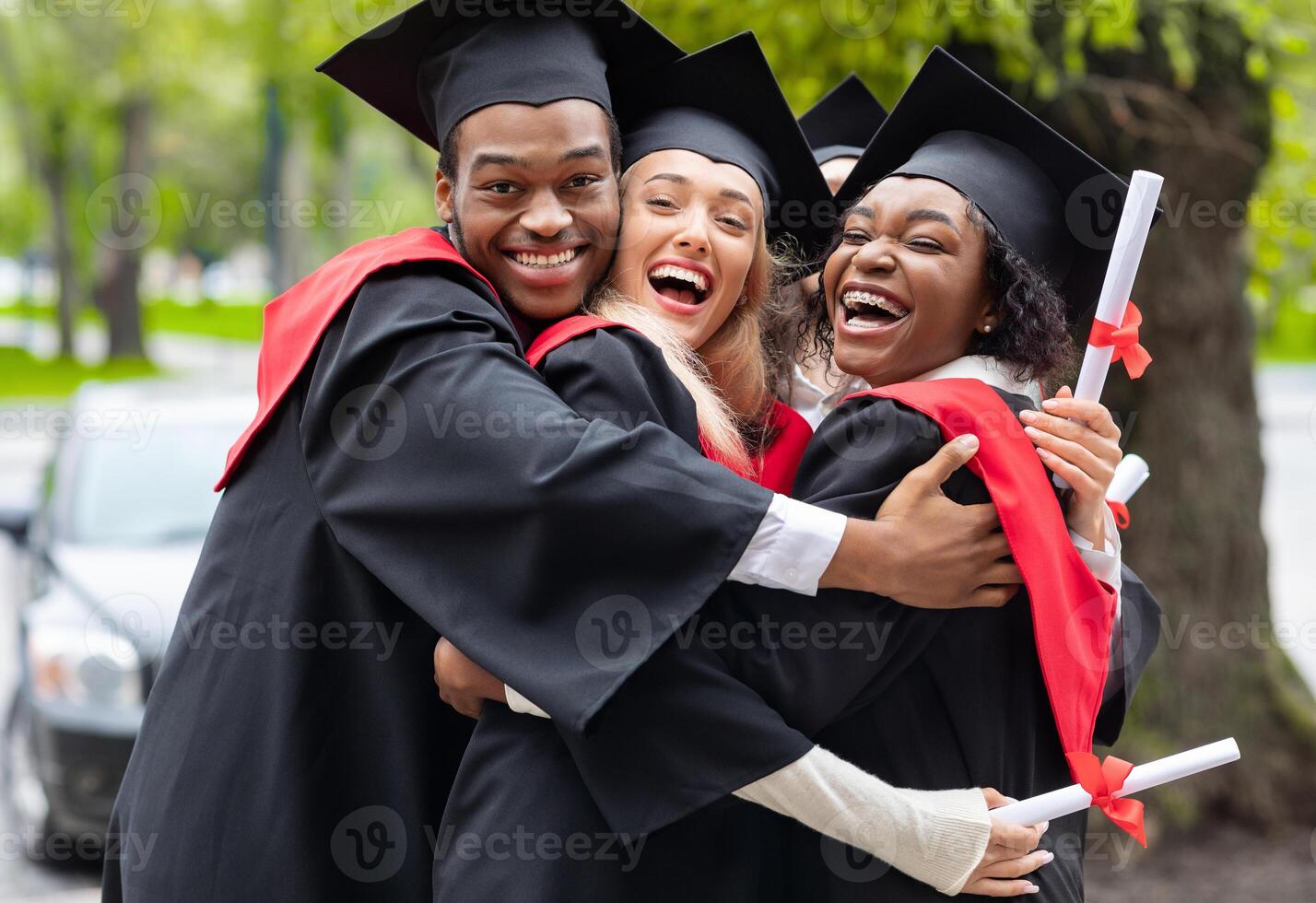 Black guy and two young ladies in graduation costumes posing at camera at university campus, holding diplomas, laughing and hugging, having happy graduation day, closeup portrait photo
