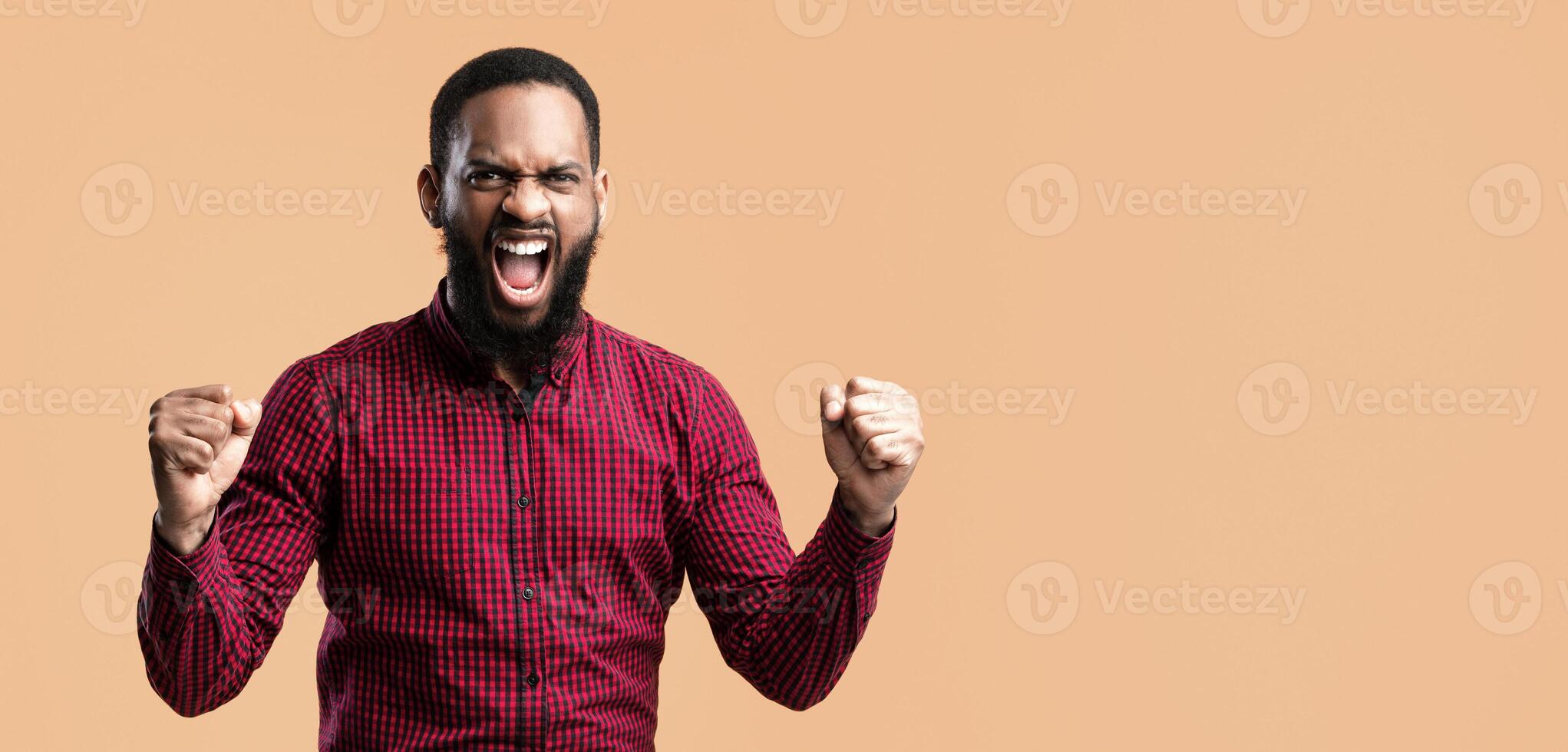 Lucky Person Concept. Studio portrait of african american guy screaming with clenched fists, isolated yellow studio background. Bearded man cheering and shouting, raising hands, celebrating victory photo