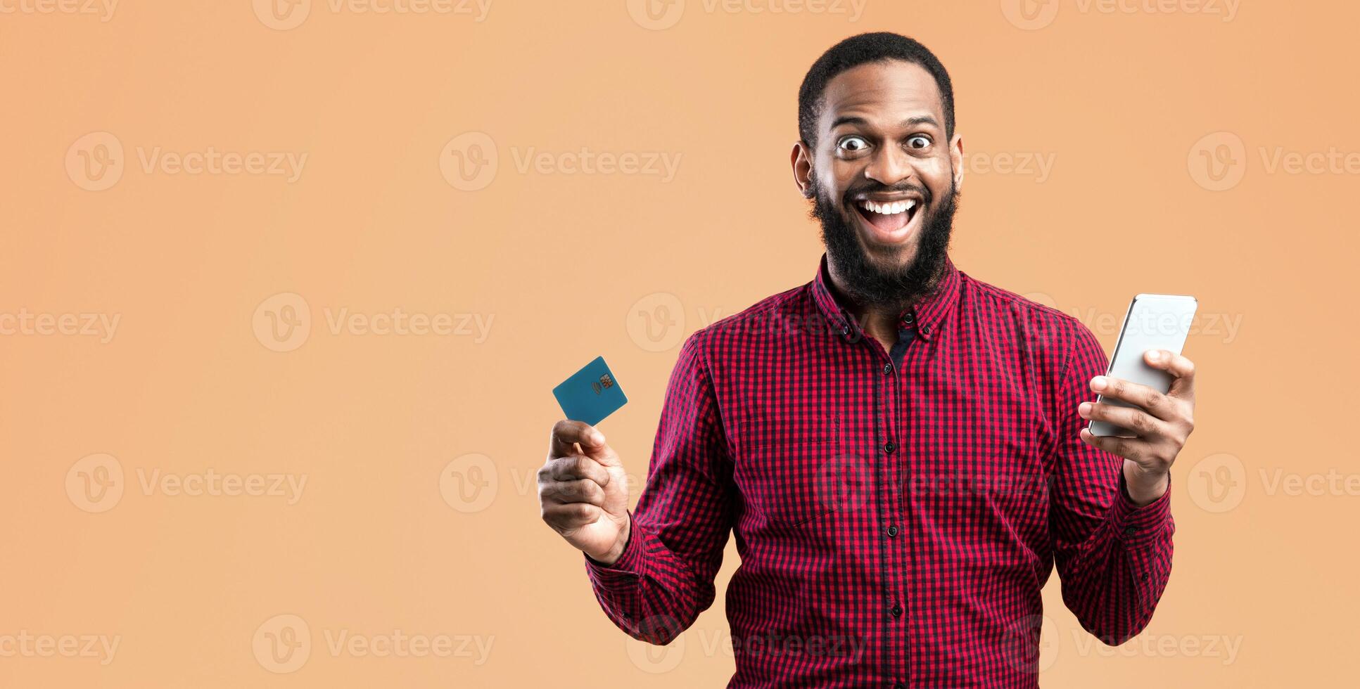 Mobile Shopping Concept. Excited African American Guy Holding Credit Card And Smartphone Posing Standing Over Yellow Background. Studio Shot. Joyful Black Man Making Online Purchase photo
