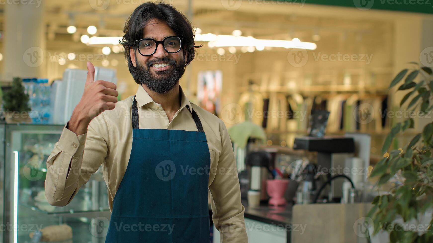 Coffee shop worker show like agree recommend gesture approve hand symbol successful happy smiling cafe waiter guy Indian Arabian male cafeteria working showing thumb up barista man small business photo