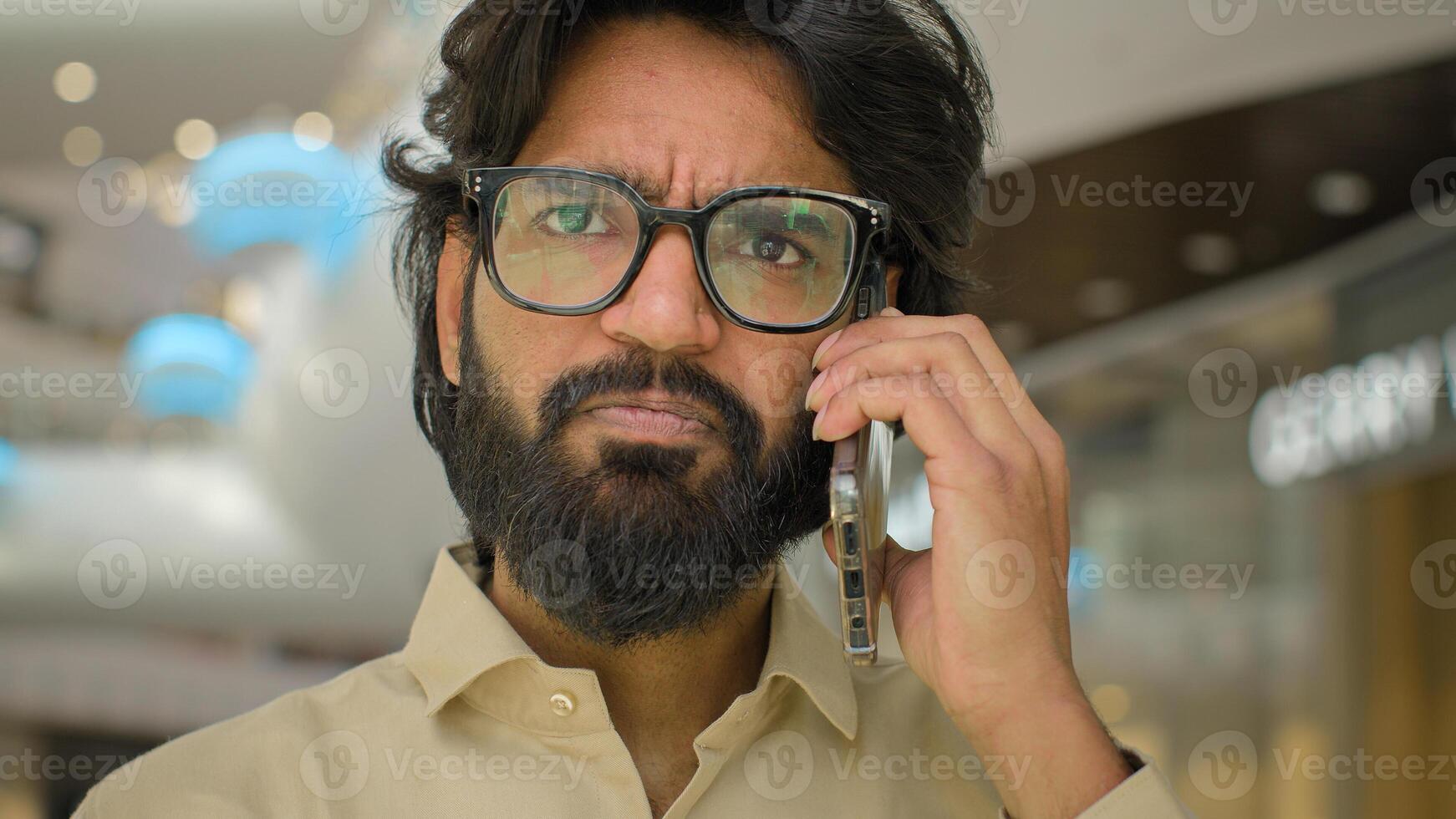 Portrait in office sad Arabian man in glasses talking phone answer business call Close up serious worried Indian male businessman executive CEO speaking distant communication talk mobile conversation photo
