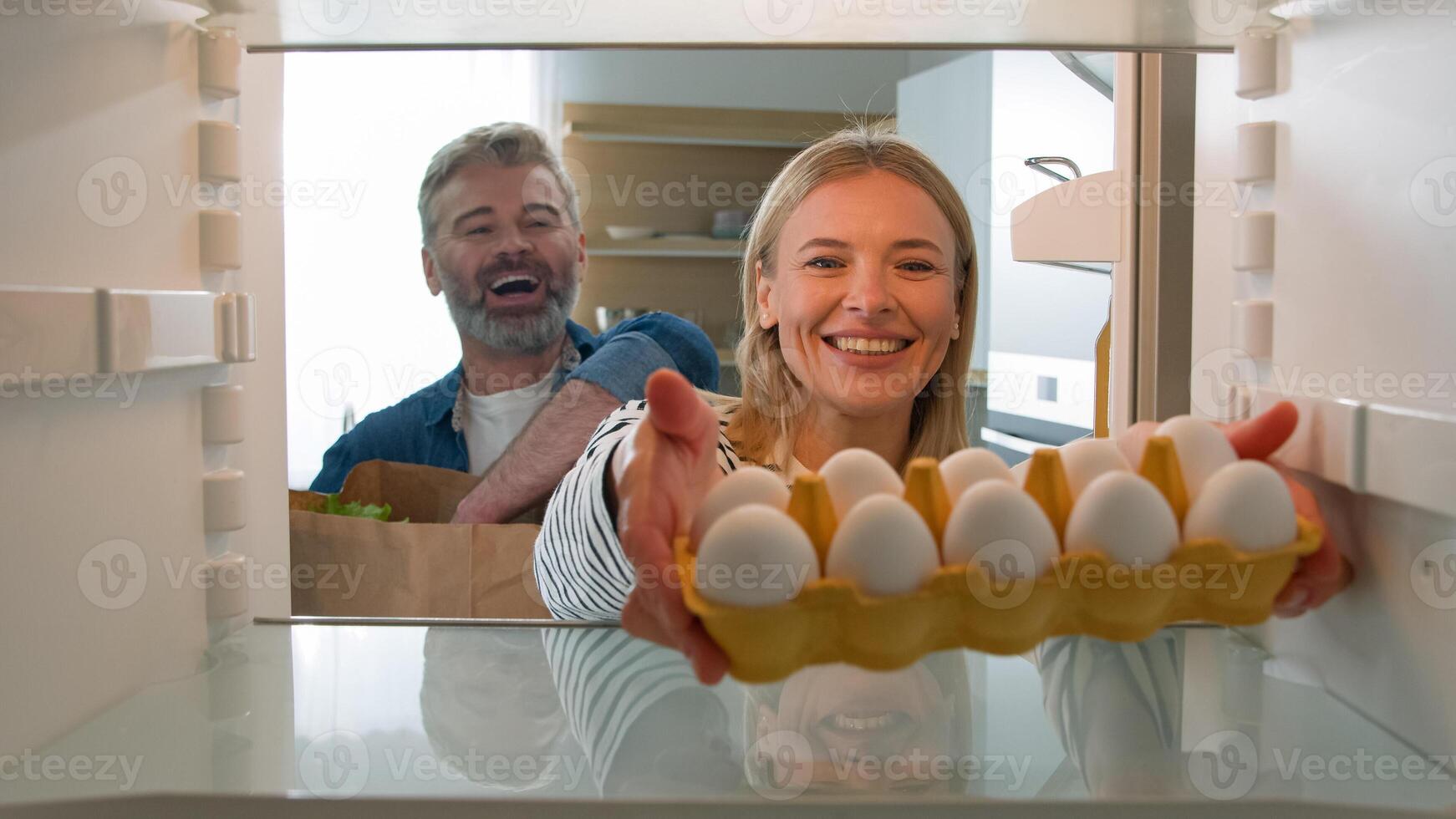 Point of view POV from inside refrigerator middle-aged couple spouses adult man and woman husband and wife taking vegetables food from paper bag putting products on fridge shelf. Healthy nutrition photo