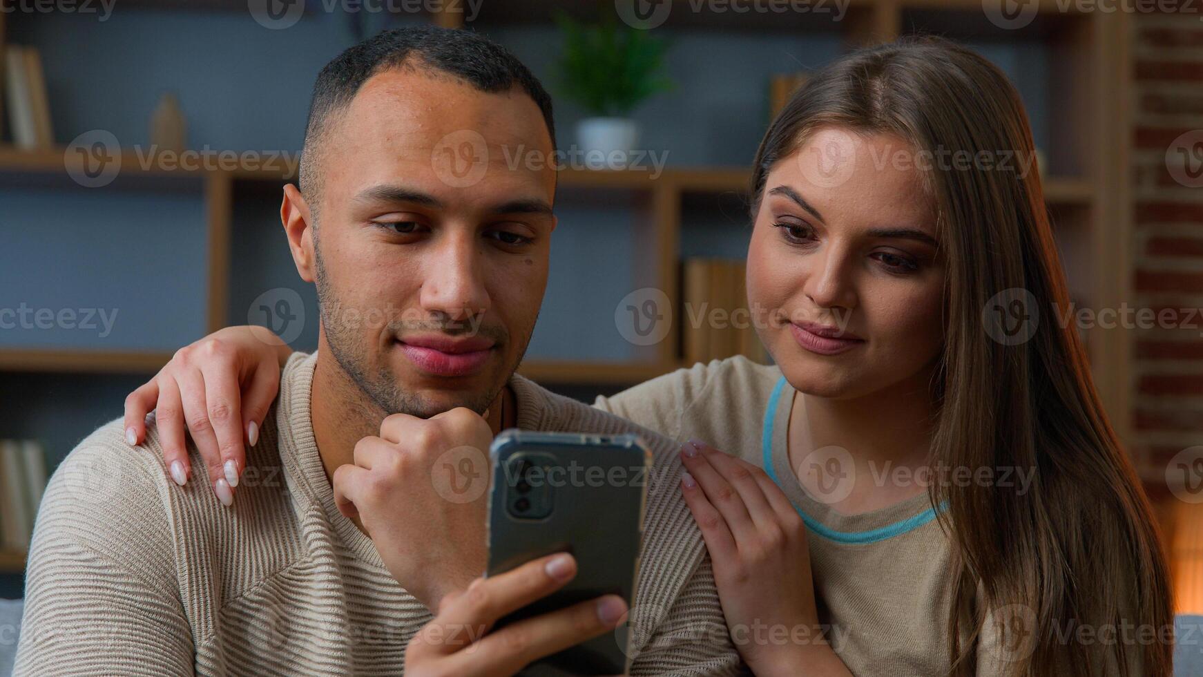 Ethnic couple use mobile phone apps smiling african american man and caucasian woman looking at smartphone screen watch online social media chatting home leisure with cellphone internet shopping photo