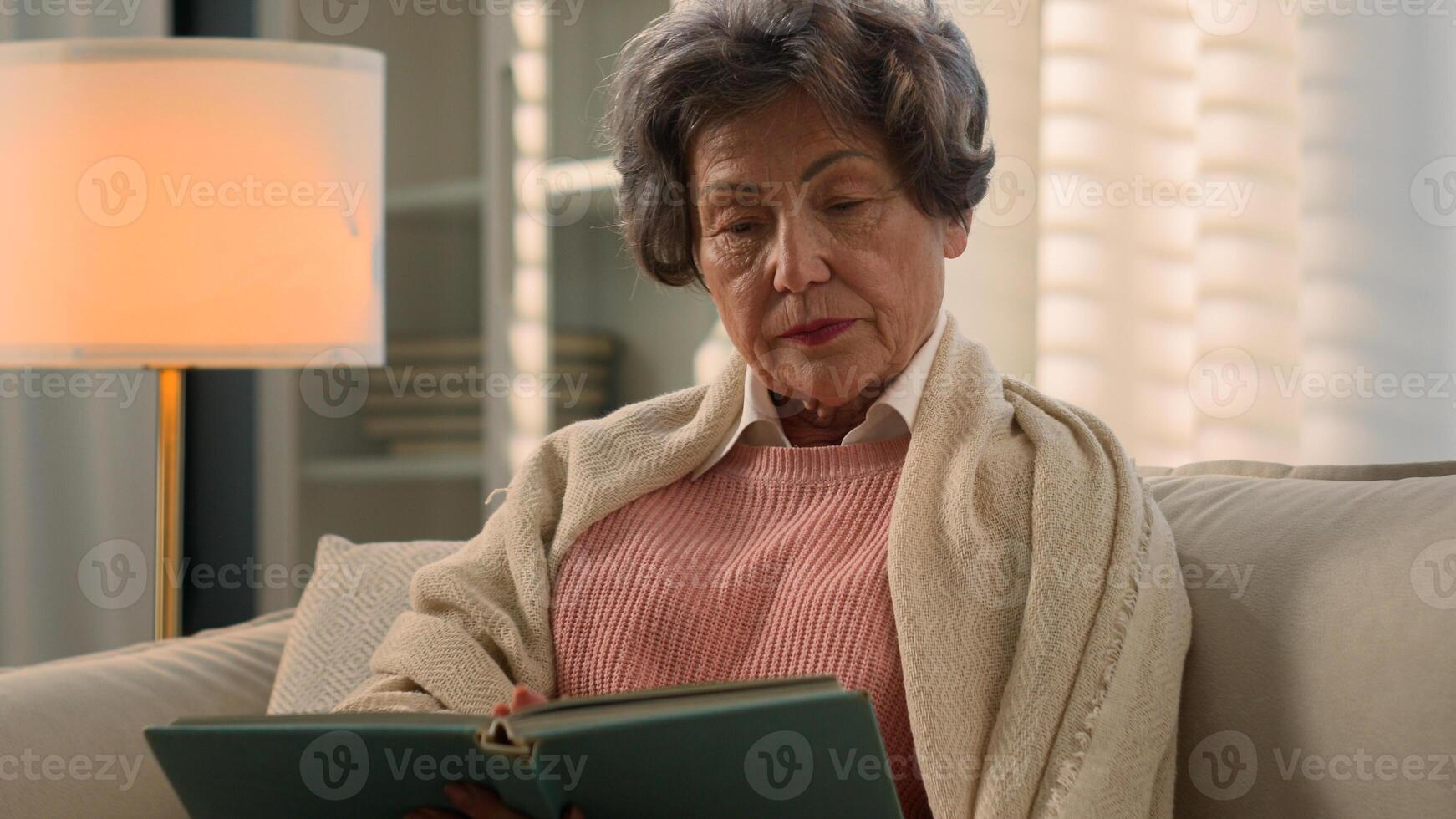 Serious dissatisfied Caucasian old woman senior mature lady covered with blanket calm elderly female retired middle-aged granny grandmother at home couch reading bad book enjoy read novel literature photo