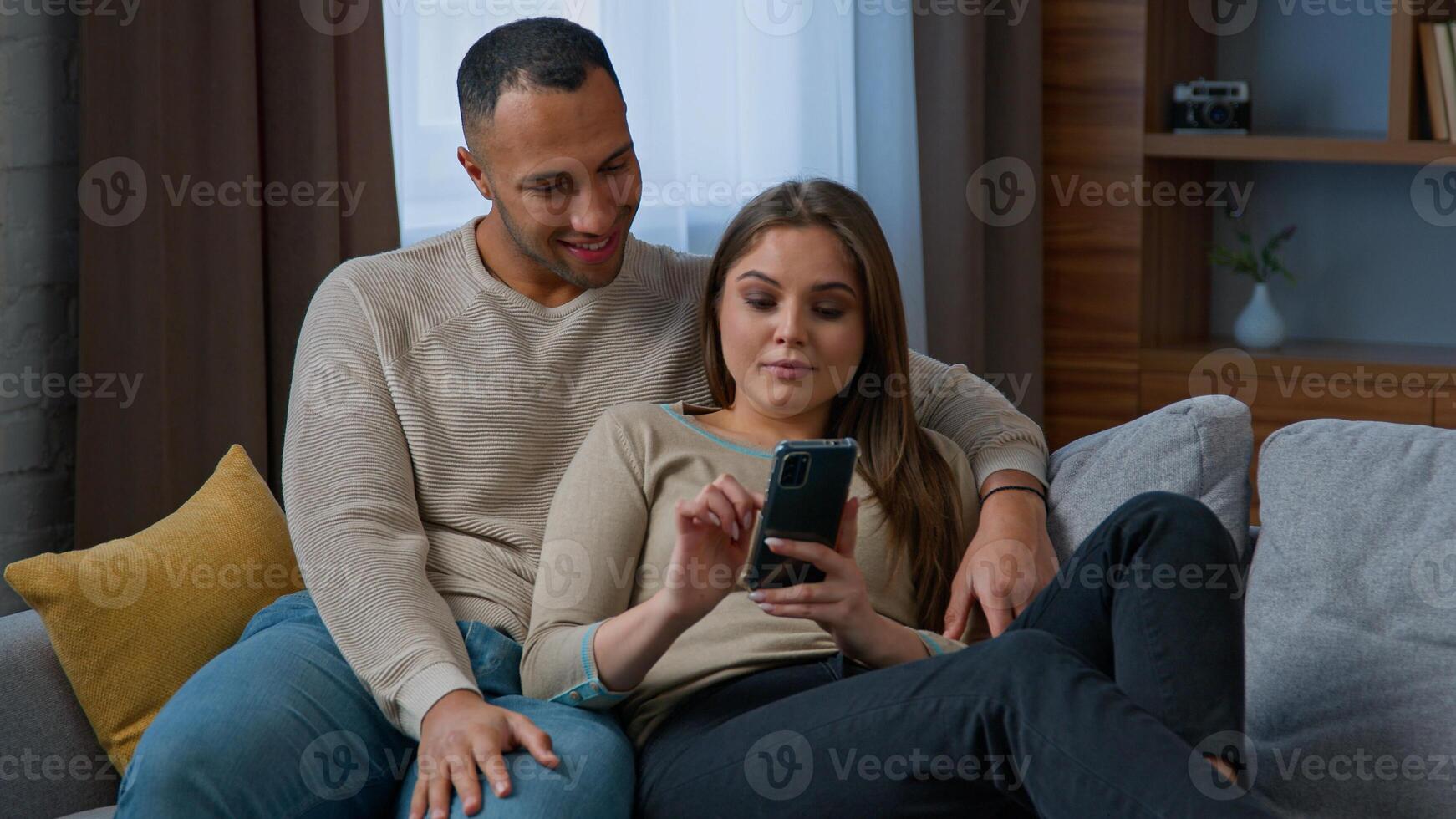 Couple multiracial hispanic woman african american man resting on cozy sofa with smartphone looking at phone screen watch photo use mobile app talking spend weekend at home with cellphone gadget