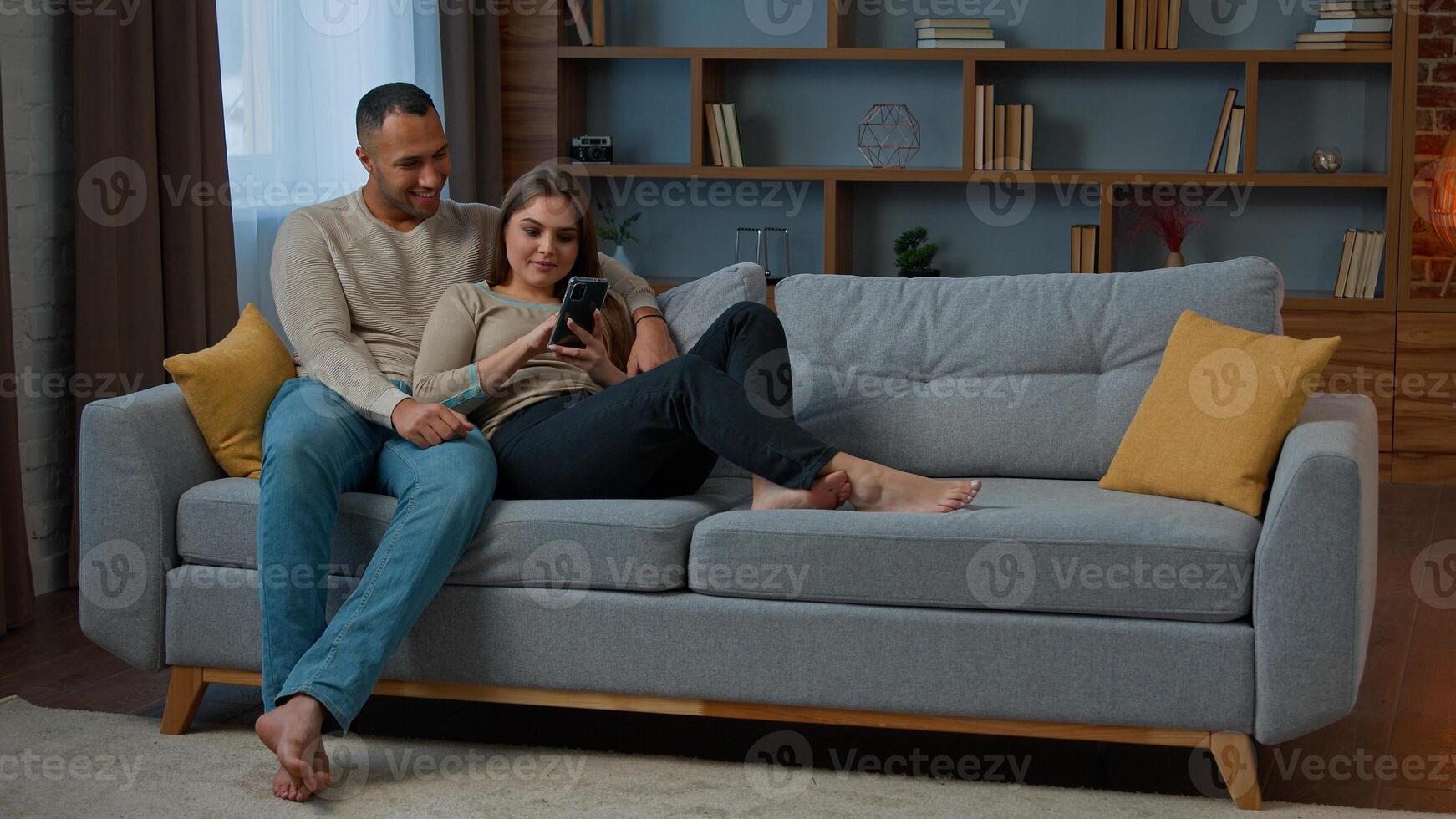 Diverse man and woman interracial couple homeowners relax on sofa talk call communicate online with mobile phone web app happy diverse wife husband buy internet goods chatting read social media photo