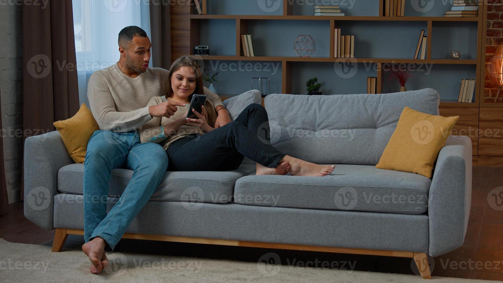 Diverse man and woman interracial couple homeowners relax on sofa talk call communicate online with mobile phone web app happy diverse wife husband buy internet goods chatting read social media photo