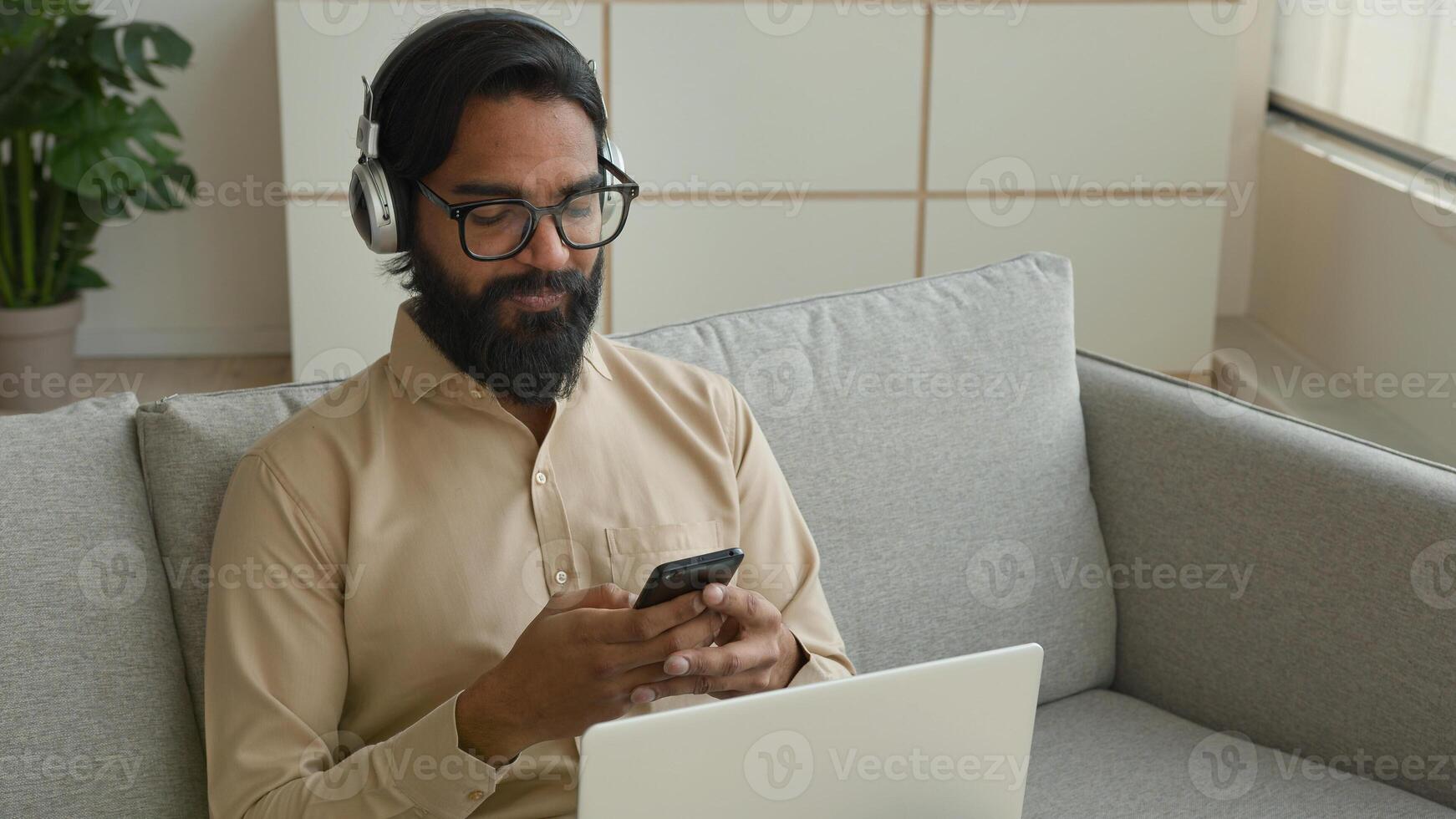 Arabian Indian businessman man multitasking working with devices at home muslim guy in headphones talk chat meeting call on laptop typing cell phone message use mobile app online communication photo