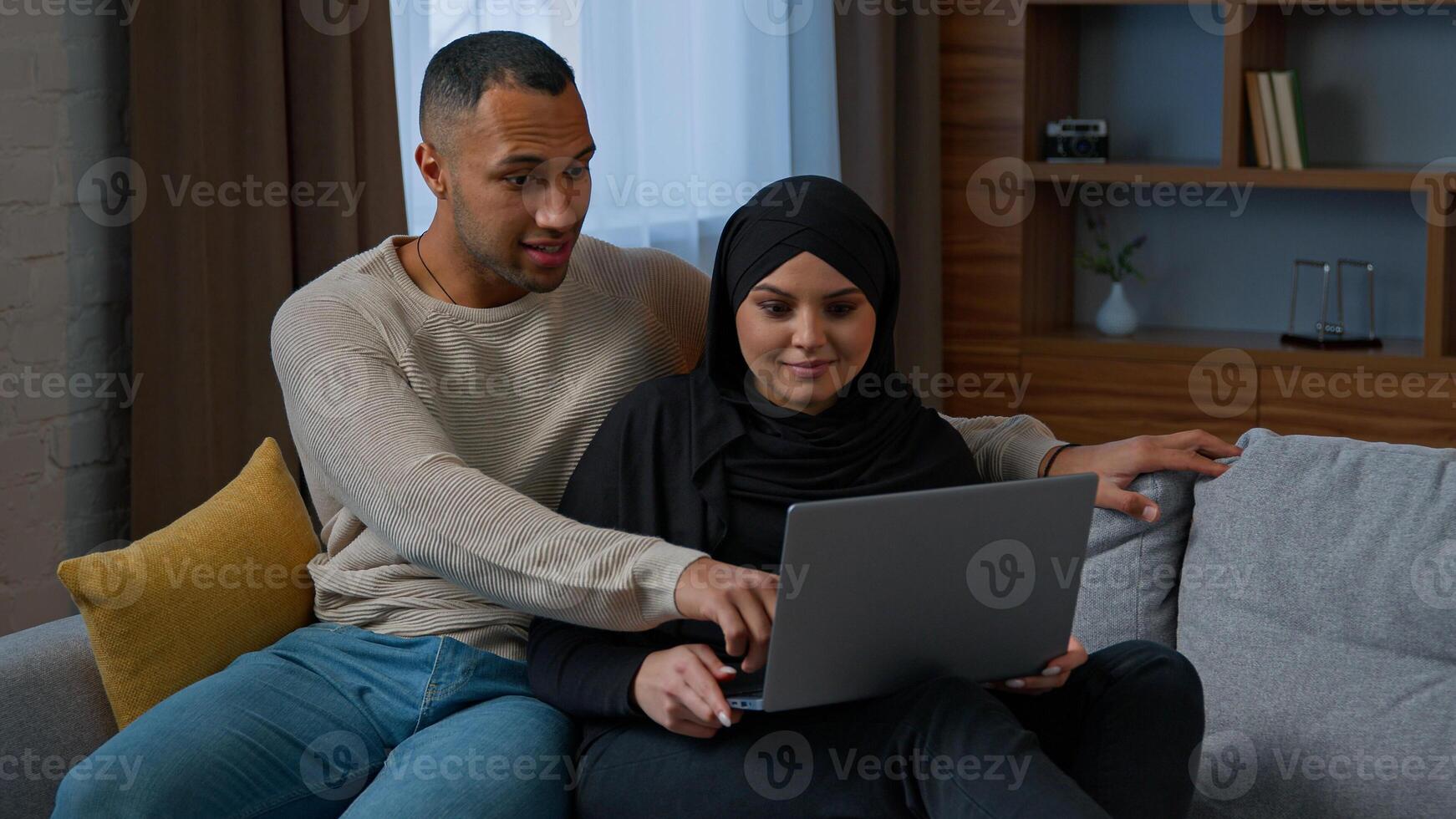 Married couple sit on sofa use laptop device choosing new apartment on internet website multiracial family shocked amazed diverse man woman search information by computer read good news talking indoor photo