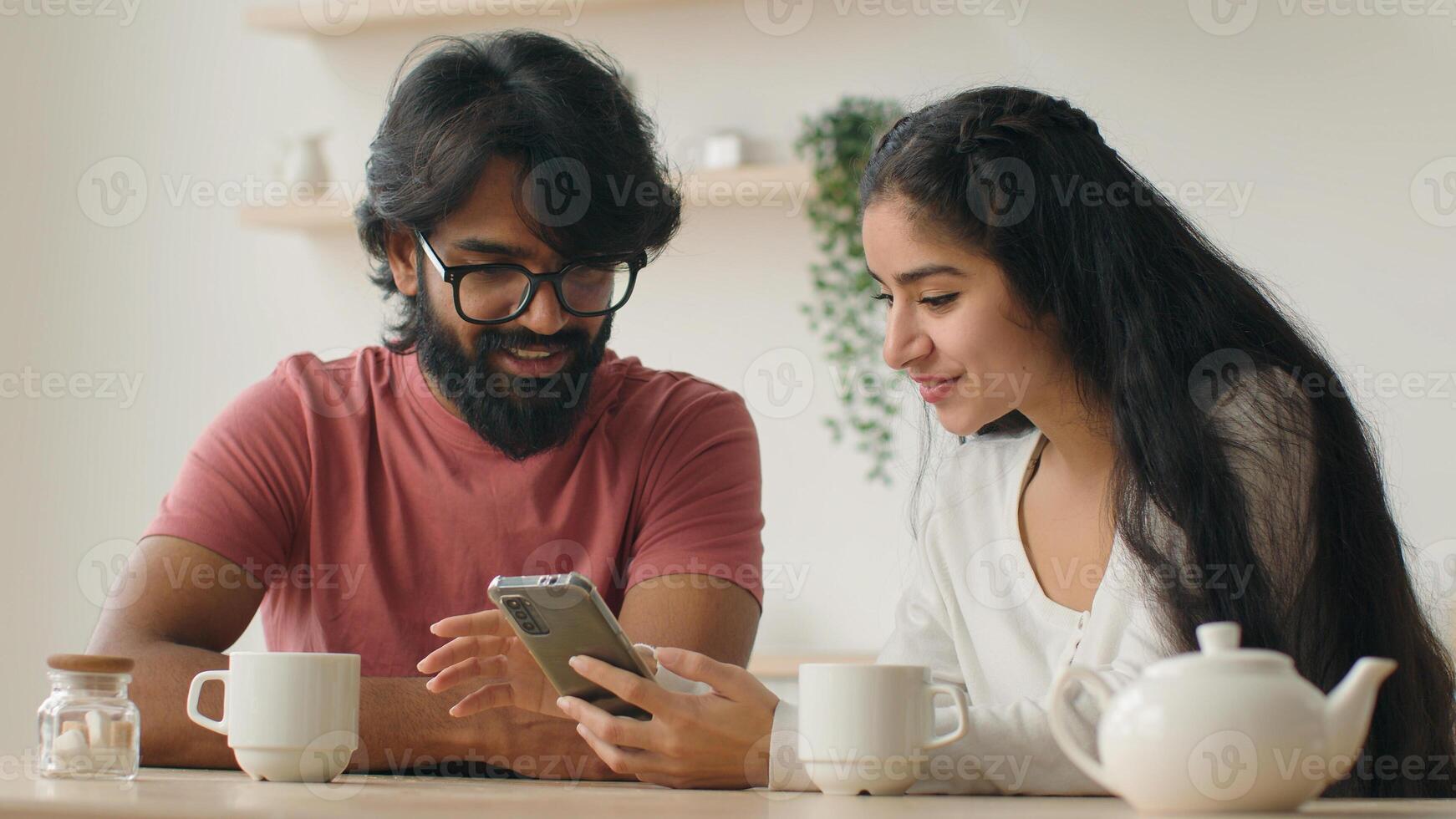 Diverse couple using smartphone arabian woman indian man in kitchen look mobile phone screen buying goods online order food delivery services internet shopping in ecommerce app talk browsing website photo
