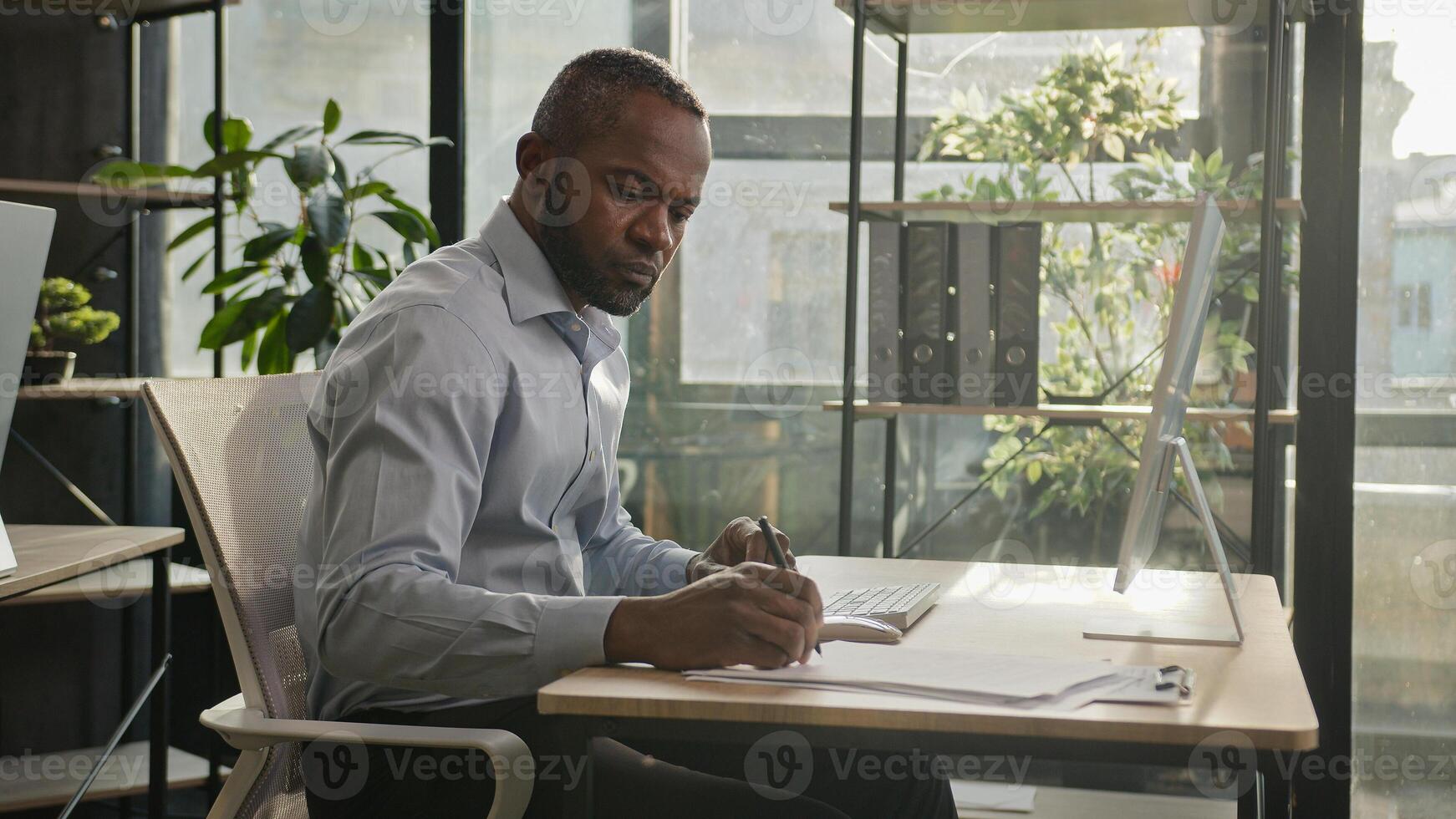African american mature senior businessman 50s ethnic man sit in sun shining sunlight office banker manager write notes business documents writing text on paper count loan bills enter data at computer photo