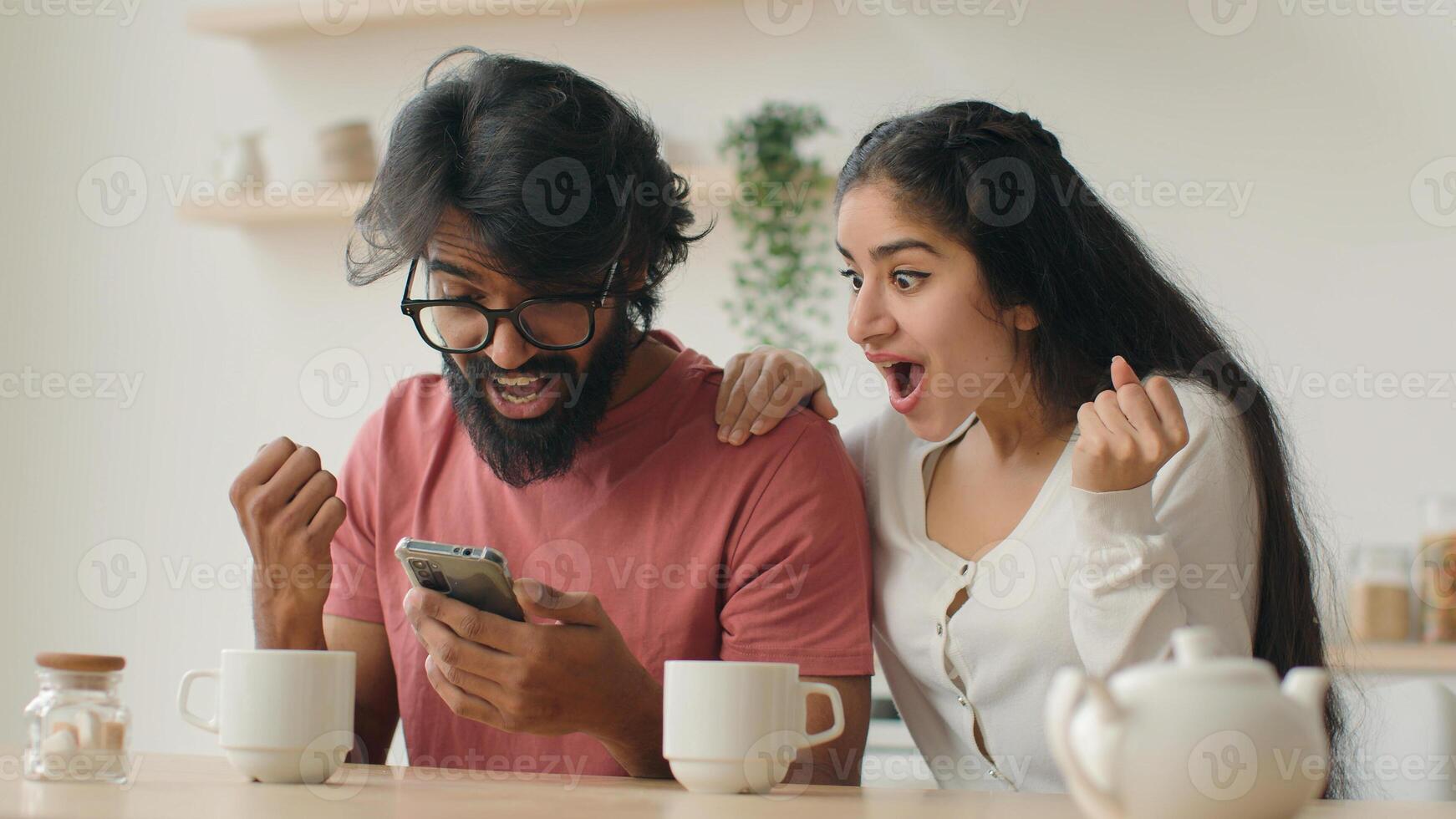 Multiracial couple indian man and woman sit in kitchen using mobile phone application enjoying web surfing read news win online bonus on smartphone happy ethnic spouses rejoice victory give high five photo