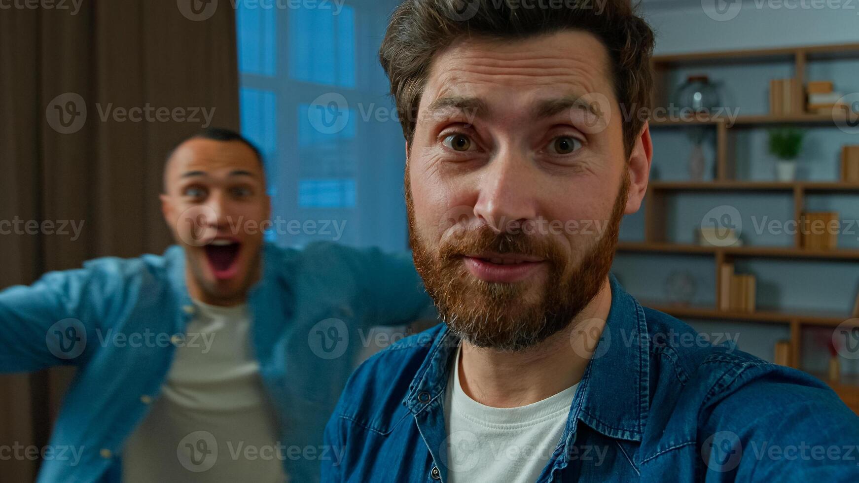 Happy caucasian man blogger vlogger 35s businessman record blog on mobile phone look at webcam camera broadcasting talk indoors on background funny african american friend running fun blogging photo