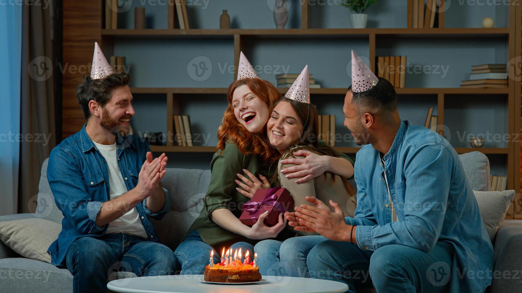 Excited woman receive gift present from friends diverse multiracial colleagues congratulate wish happy birthday presenting surprise box. Ethnic women men in party caps have fun celebrate event at home photo