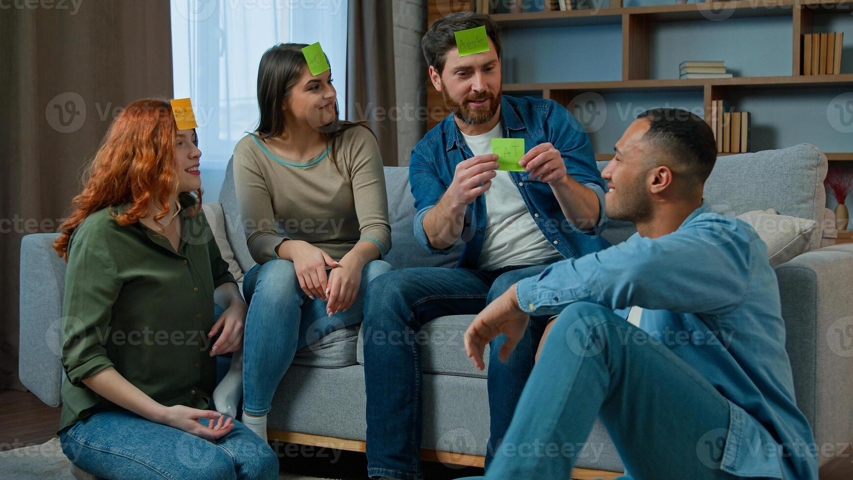 Diverse interracial friends women men playing guessing game with sticky notes on forehead play charades asking questions funny entertainment at home african american ethnic man winner win gaming level photo