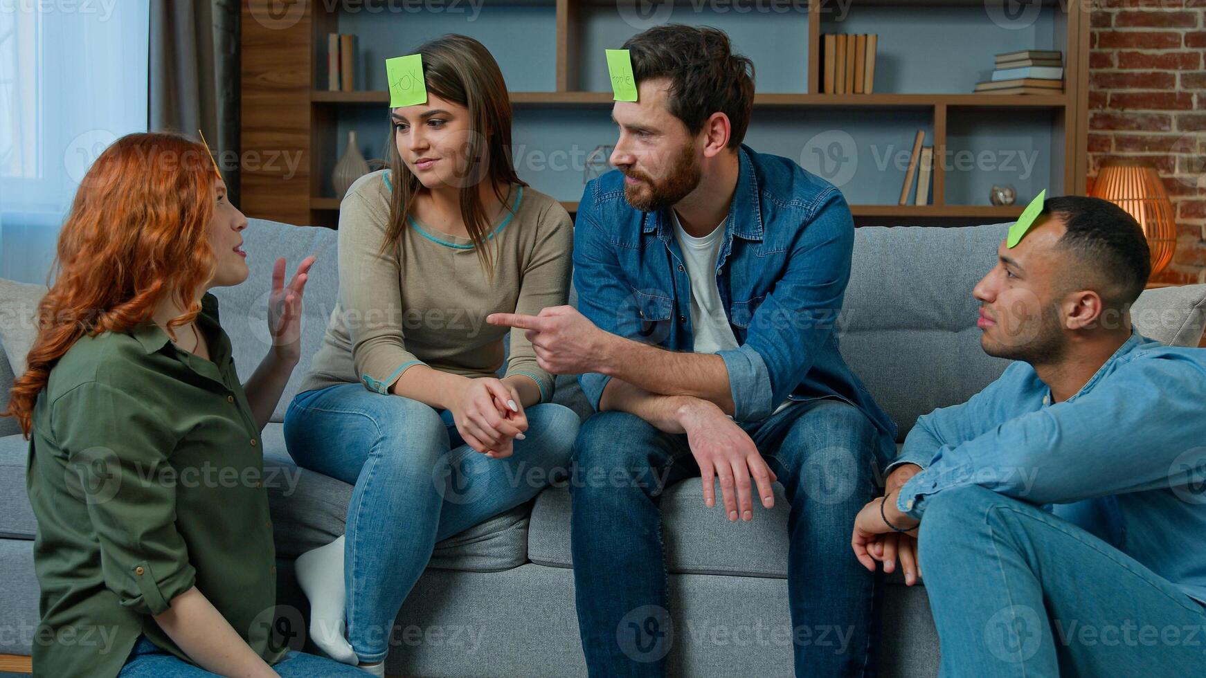 Group of multiethnic friends play charades game at home happy laughing diverse ethnic men and women guessing guess personage playing with sticky notes on head talk ask questions laugh entertainment photo
