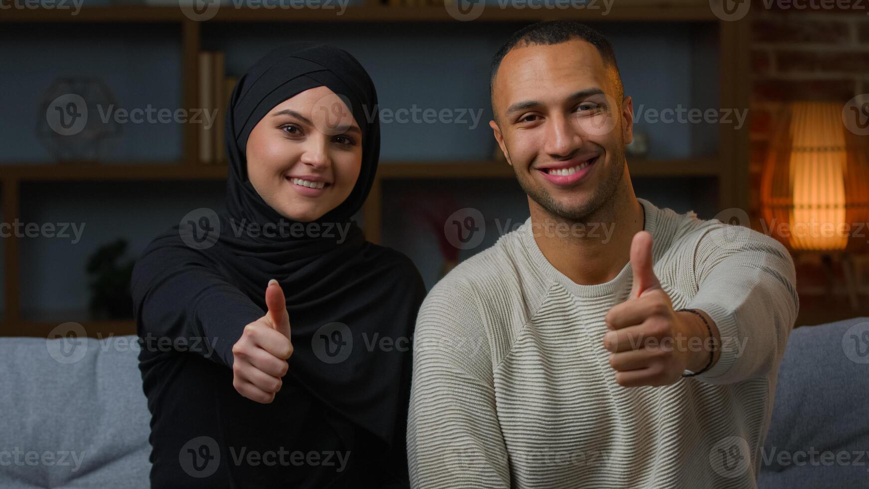 Positive interracial african american muslim couple newlyweds ethnic family homeowners woman and man sit at home showing thumb up hand gesture approve like recommend house mortgage confirm good choice photo