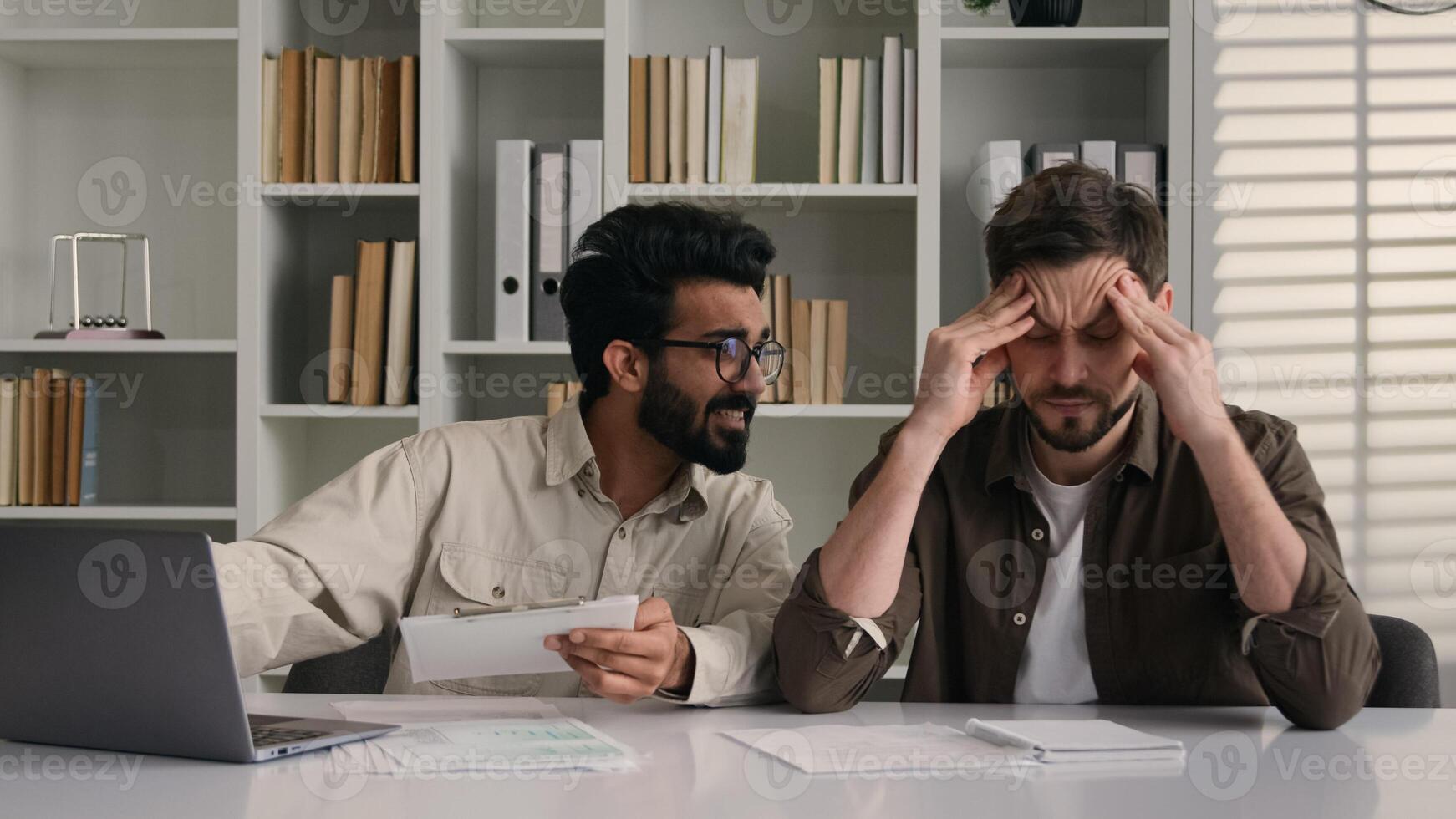 Overworked tired sick man suffer headache ignoring obsessive annoying colleague disturb talk smile discuss project with laptop two multiethnic businessmen diverse men solve teamwork problem in office photo