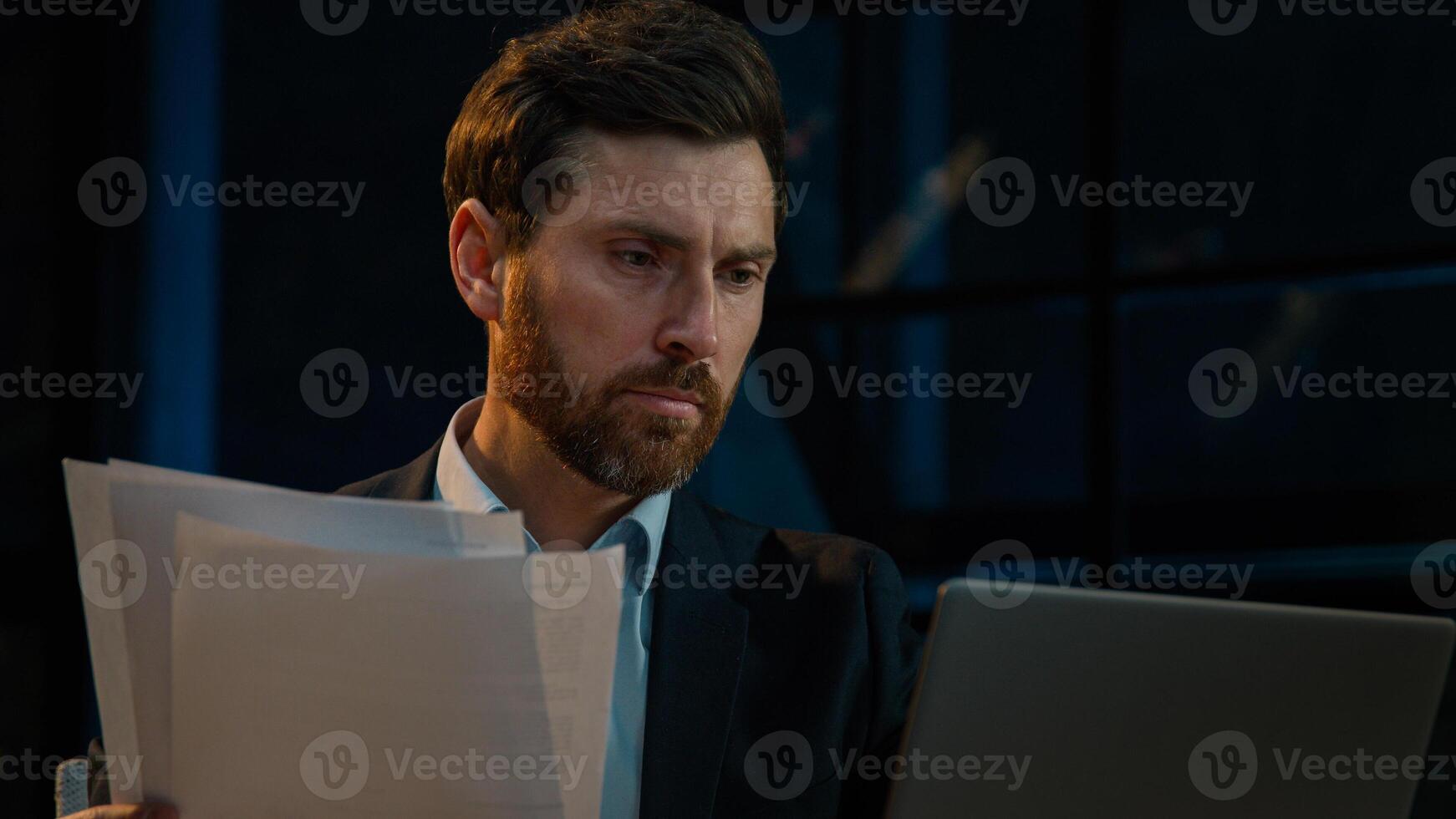 Caucasian 40s businessman employee auditor middle aged man sit in evening night office read business documents papers legal contracts pensive employee think check data in computer search information photo