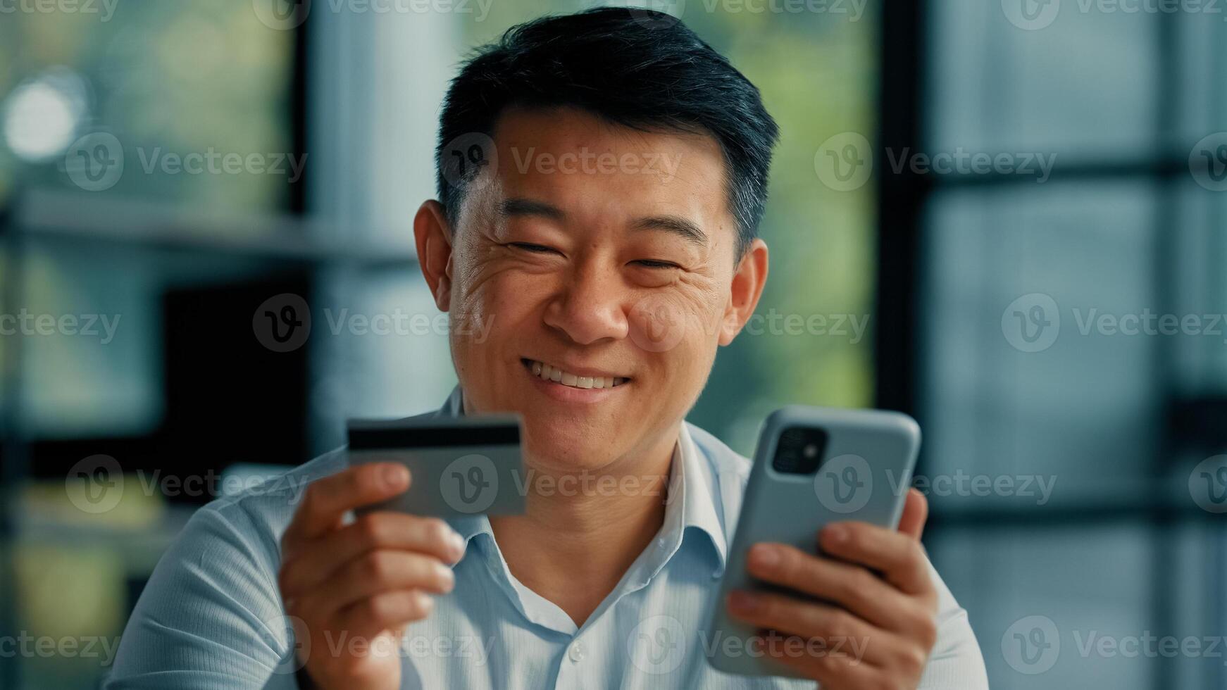 Happy Asian man client buyer businessman use banking mobile phone app at office hold plastic credit card enter secure code buy goods in internet store shopping online cryptocurrency money transaction photo