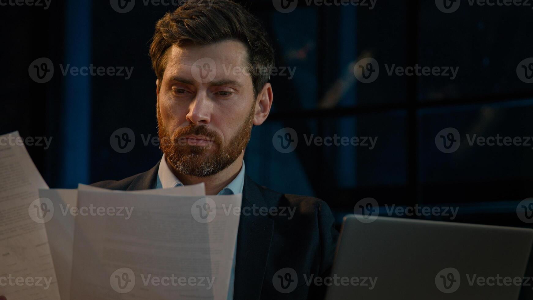 Caucasian 40s businessman employee auditor middle aged man sit in evening night office read business documents papers legal contracts pensive employee think check data in computer search information photo