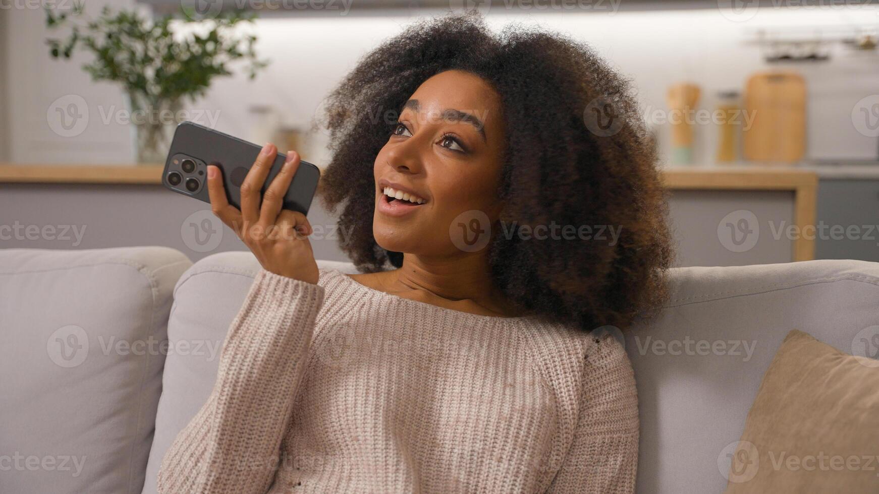 Happy African American woman on couch ethnic smiling female girl mobile phone send audio message using cell speakerphone convenient usage smartphone record voice mail recording chat voicemail at home photo