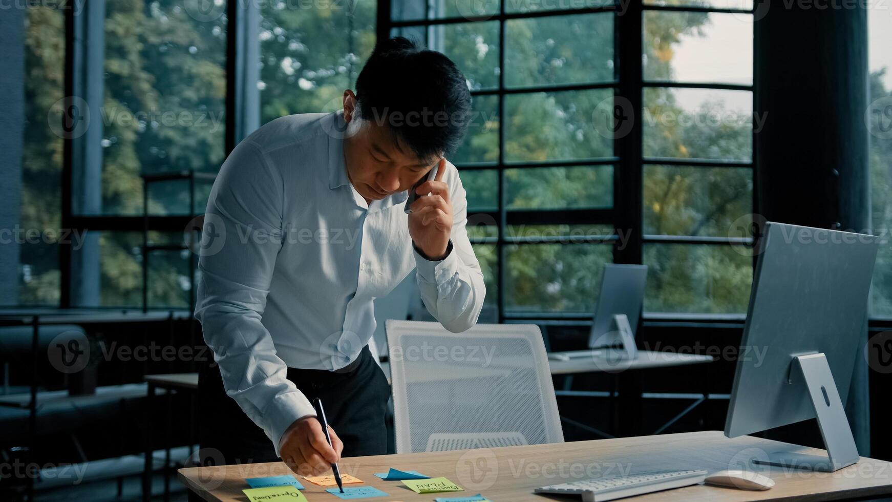 Busy asian businessman korean man working stand at table in modern office talk on phone give professional consultation by mobile call write tasks on sticky notes project paperwork multitasking concept photo