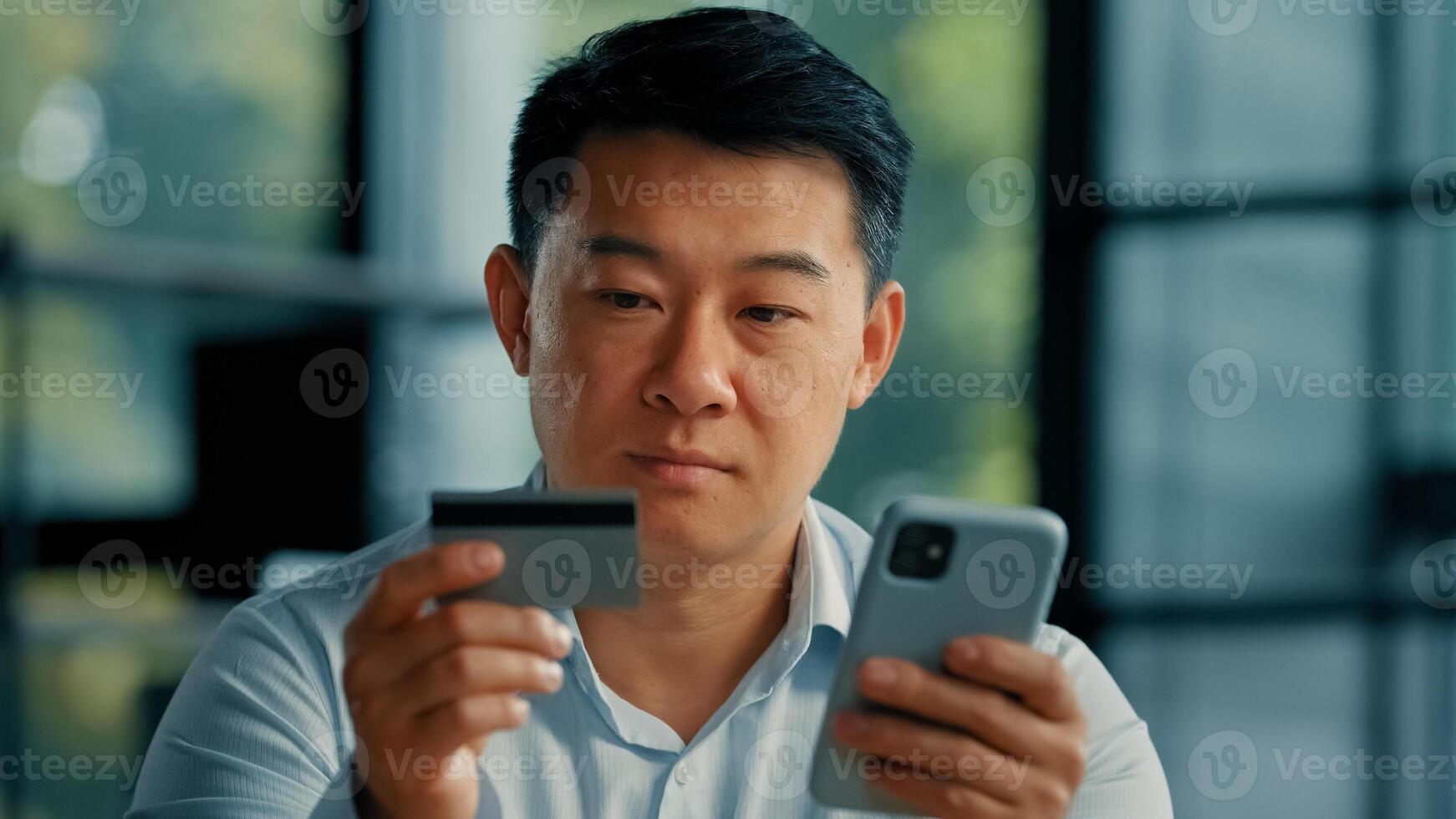 Happy Asian man client buyer businessman use banking mobile phone app at office hold plastic credit card enter secure code buy goods in internet store shopping online cryptocurrency money transaction photo