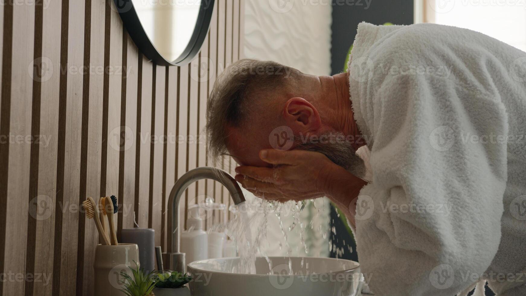Old Caucasian middle-aged senior male retired mature bearded man gray haired pensioner in bathrobe in morning hygiene routine bath sink washing face with cold water moisturizing refresh wash skin care photo