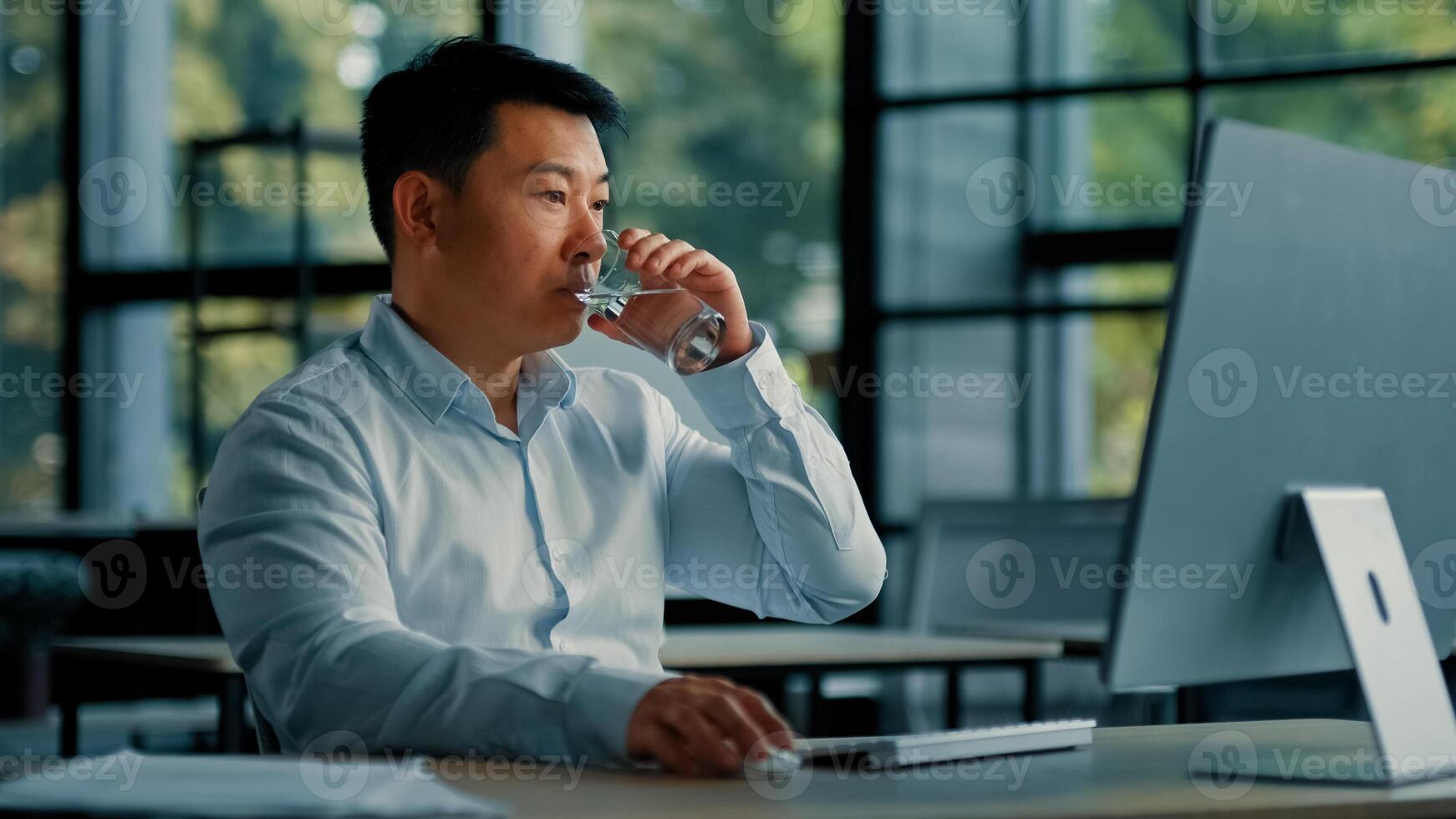 Asian korean man drink glass of cold fresh water keep health balance rejuvenation energy healthcare habit businessman sit at office work on computer make pause quench thirst body hydration bodycare photo