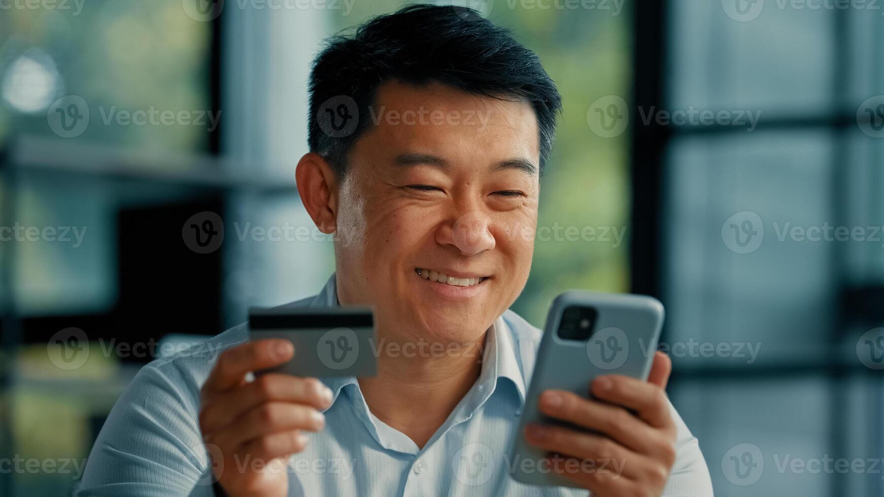 Happy Asian man client buyer businessman use banking mobile phone app at office hold plastic credit card enter secure code buy goods in internet store shopping online cryptocurrency money transaction photo