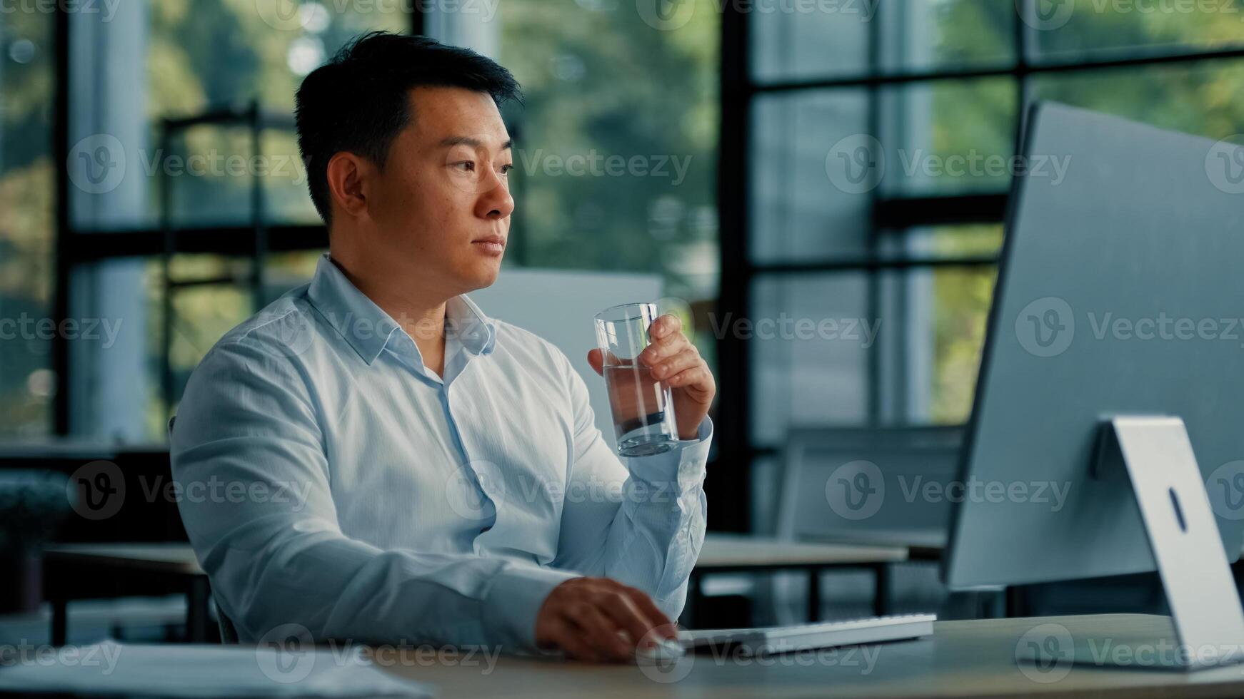 Asian korean man drink glass of cold fresh water keep health balance rejuvenation energy healthcare habit businessman sit at office work on computer make pause quench thirst body hydration bodycare photo