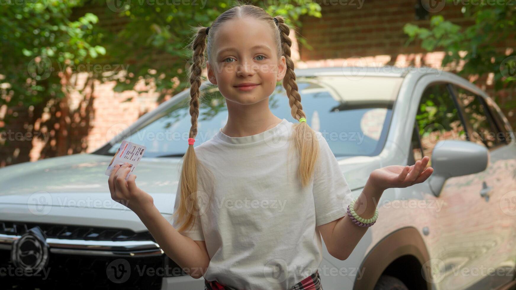 Funny confused adorable Caucasian little girl child kid smiling unsure schoolgirl looking camera showing driving license car rental service automobile what shrugging shoulders gesture puzzled at city photo